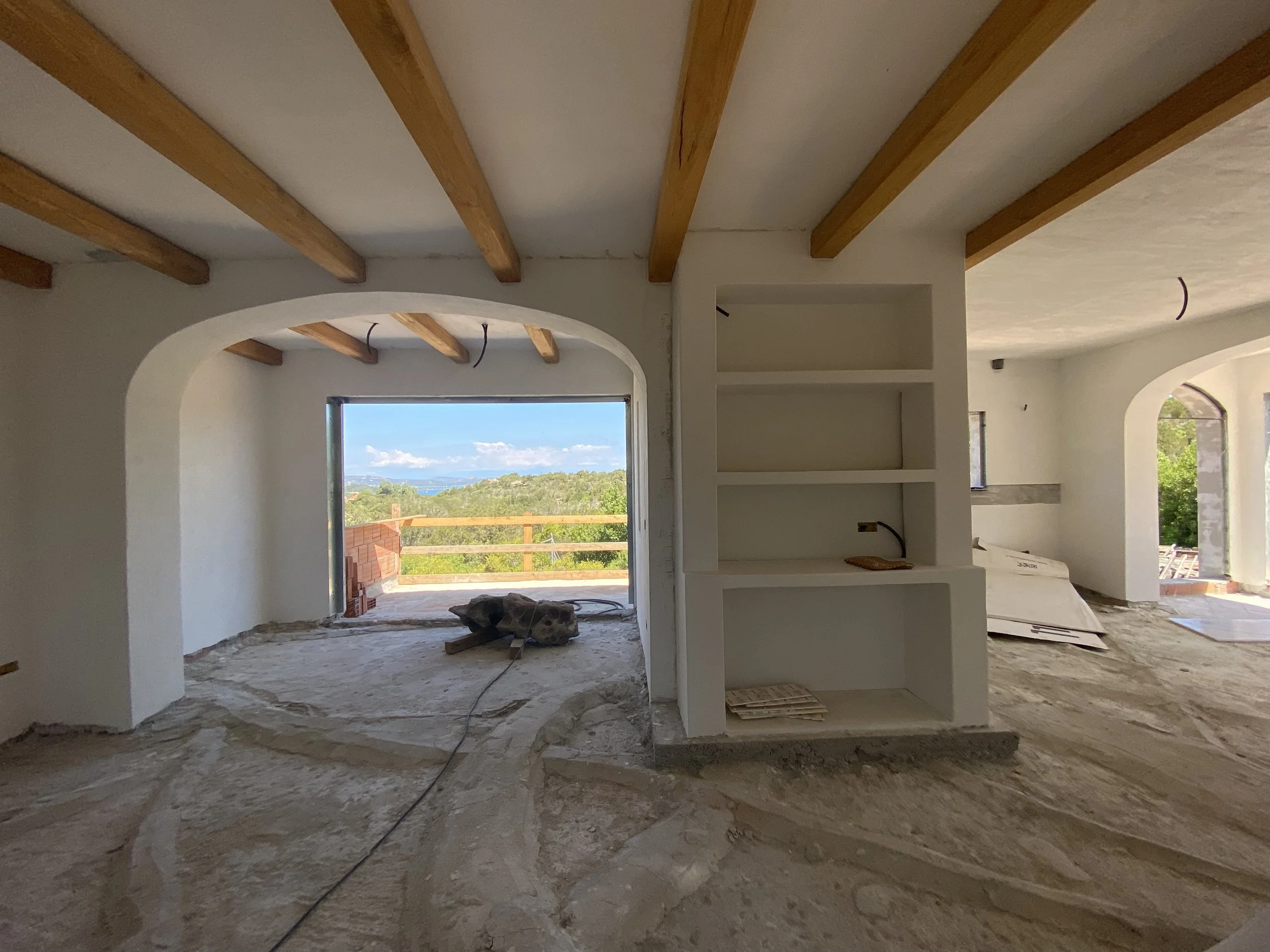 Interior of a house under construction with unfinished flooring, white walls, wooden ceiling beams, and large windows with scenic views.