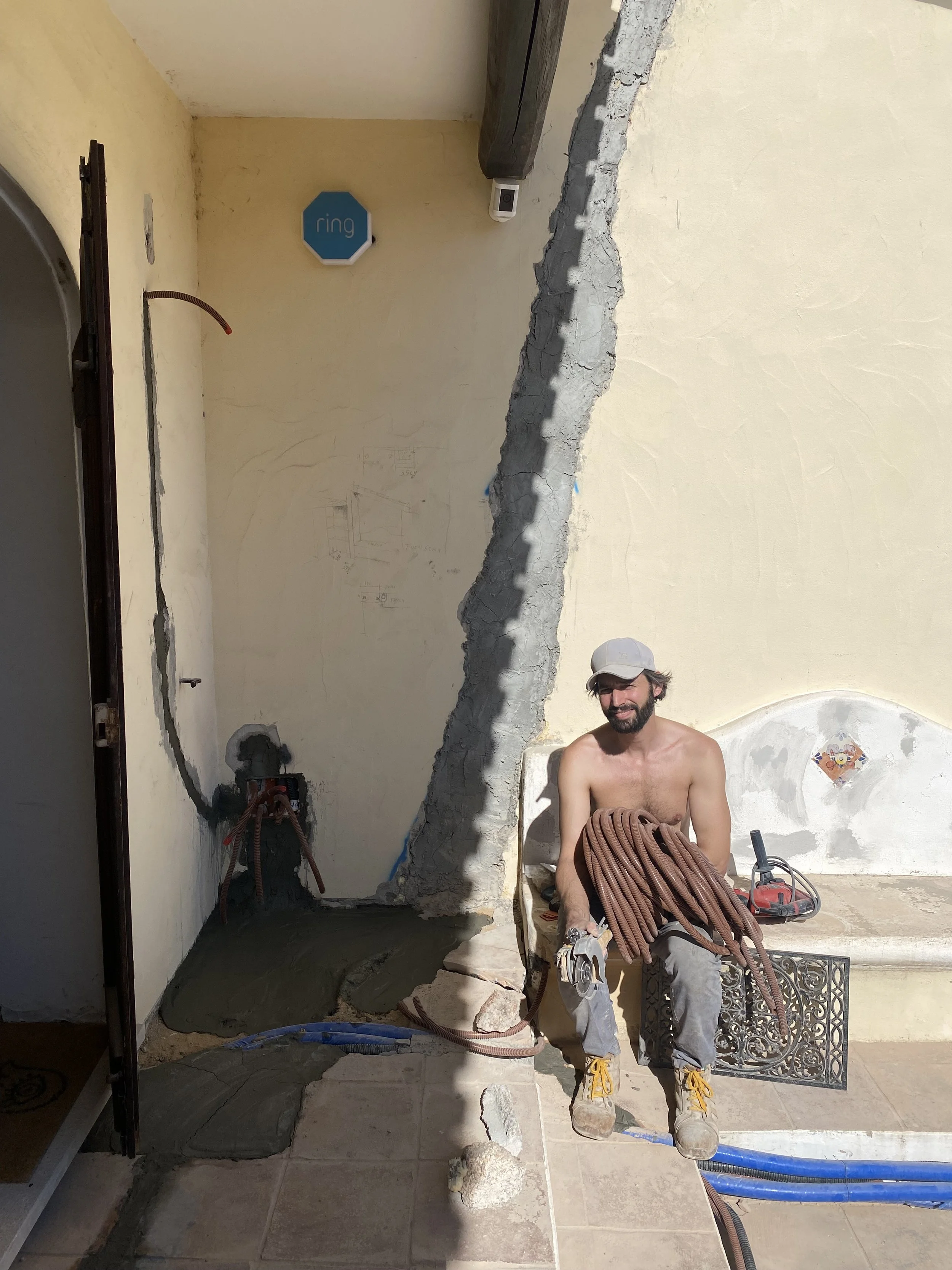 A shirtless construction worker sitting on a bench, holding a lot of coiled brown hoses, with construction tools, set against a weathered yellow wall with a broken section and a decorative ceramic tile, outdoors on a sunny day.