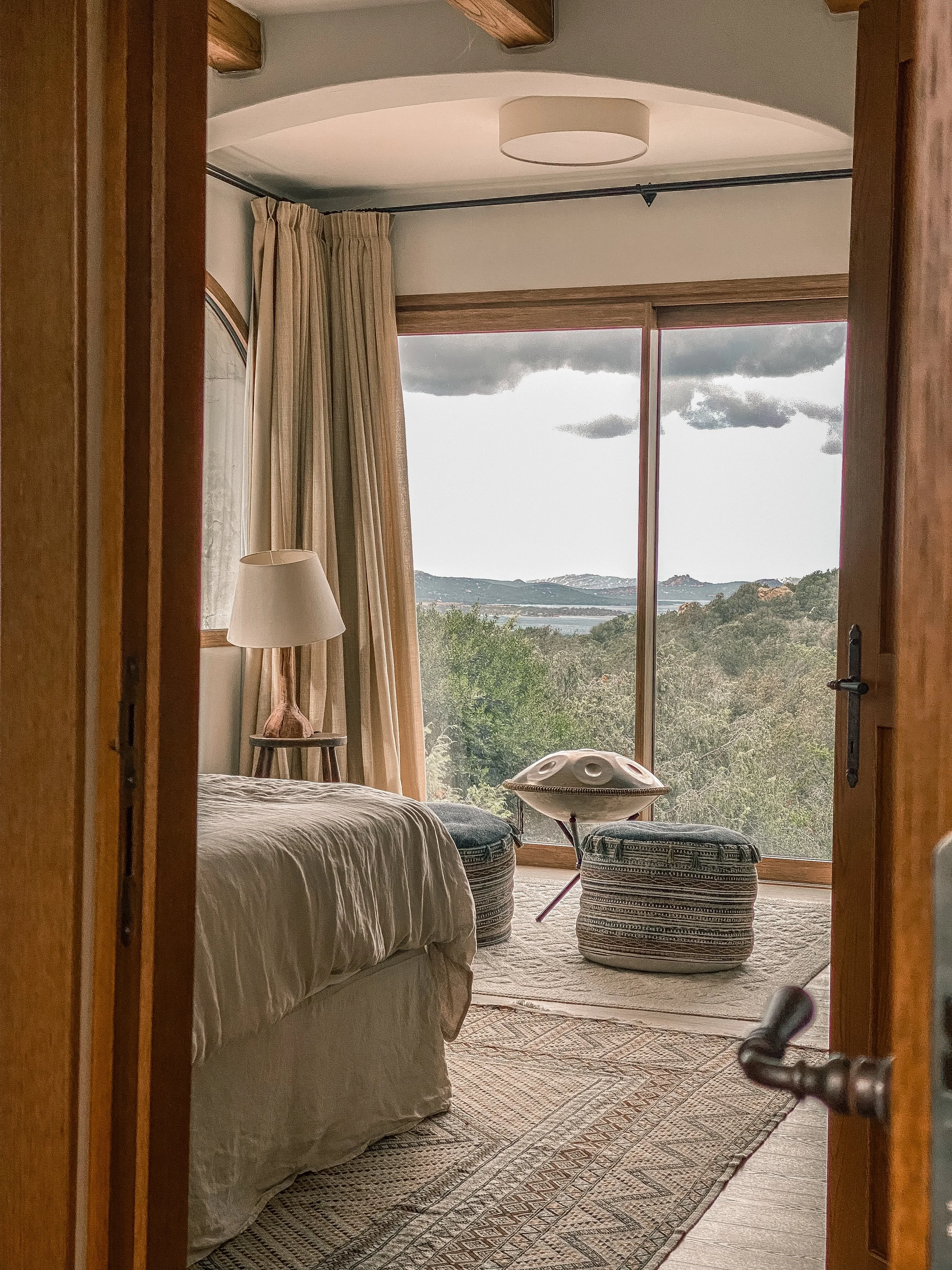 View from a hotel room looking out a large glass sliding door to a landscape with trees, hills, and cloudy sky. Inside, part of a bed, a bedside lamp, two patterned ottomans, and a musical instrument resembling a handpan are visible.