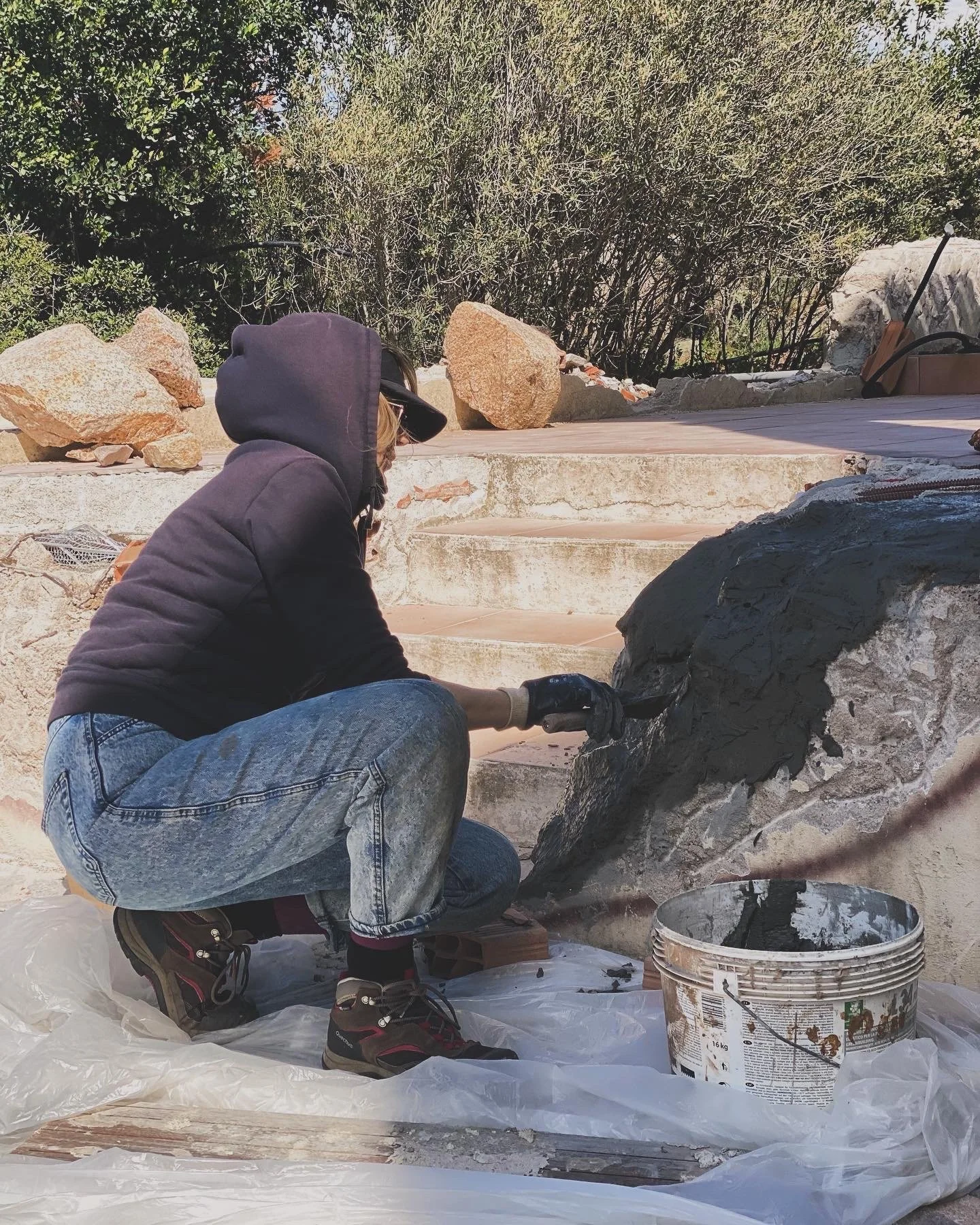 Person wearing a hoodie, gloves, and jeans applying black cement to a stone surface on an outdoor project with rocks, plants, and tools in the background.