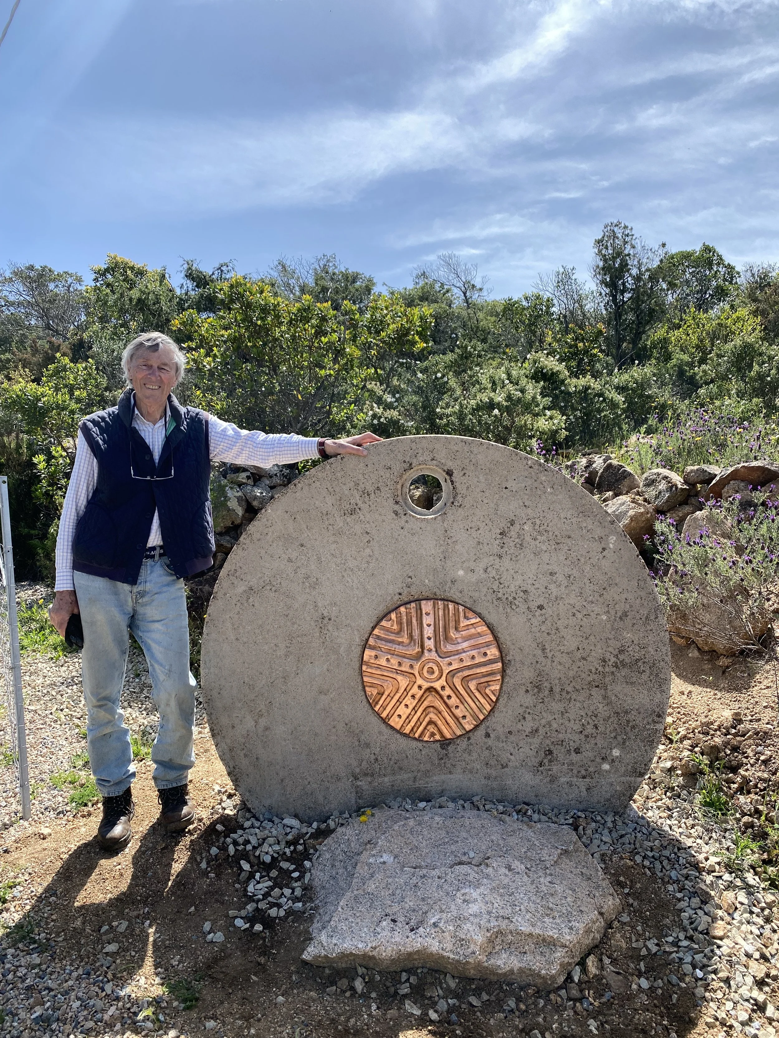 A woman standing outdoors beside a large stone monument with a copper-colored circular design in the center. She is smiling, wearing a white shirt, a dark vest, jeans, and boots. The background includes green bushes, rocks, and a partly cloudy sky.