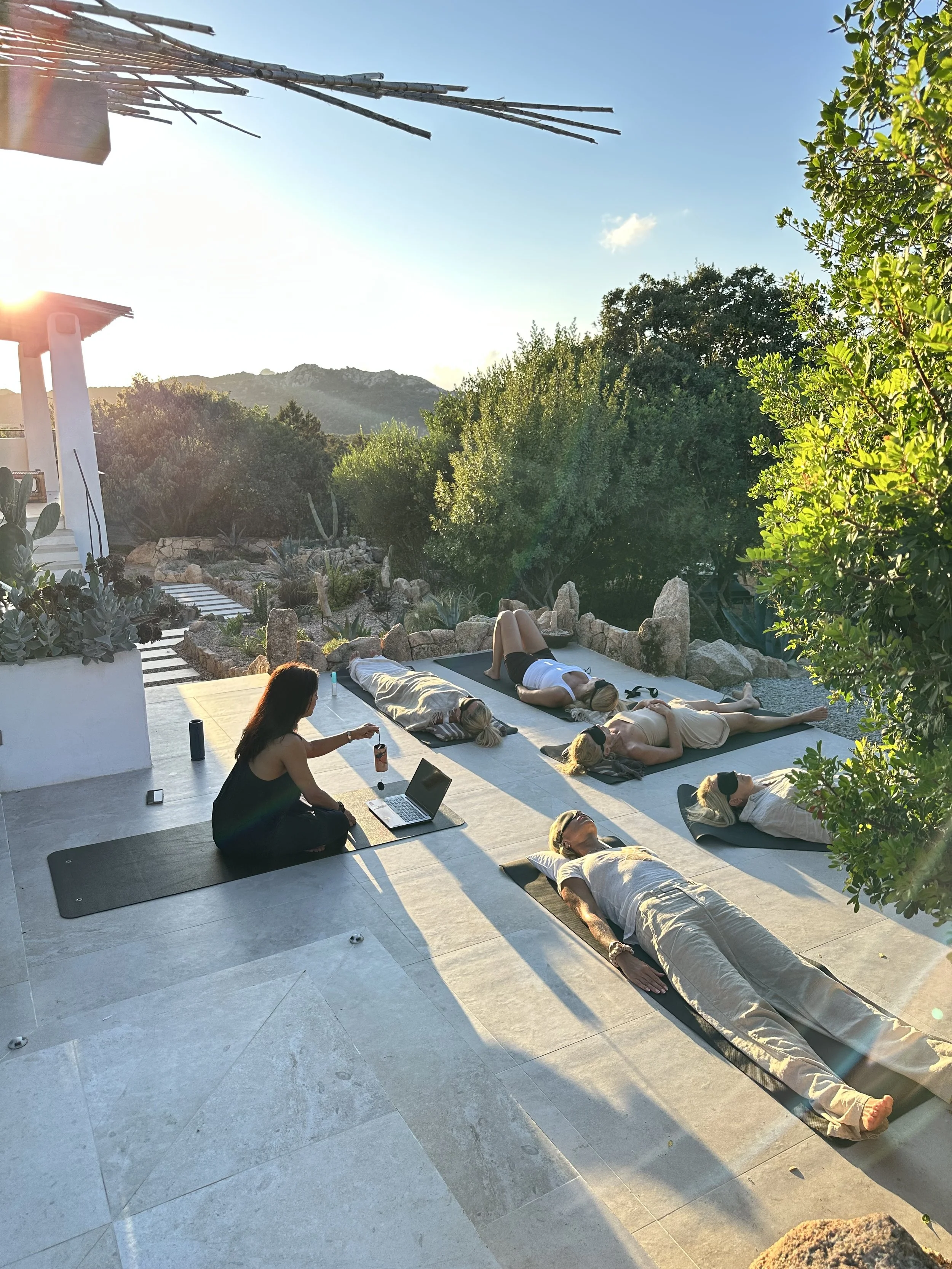 A person receiving a chiropractic treatment outdoors on a blue massage table, next to a woman resting on the table in a private villa in Sardinia. The setting is a rocky landscape with large boulders, greenery, and a small waterfall. 