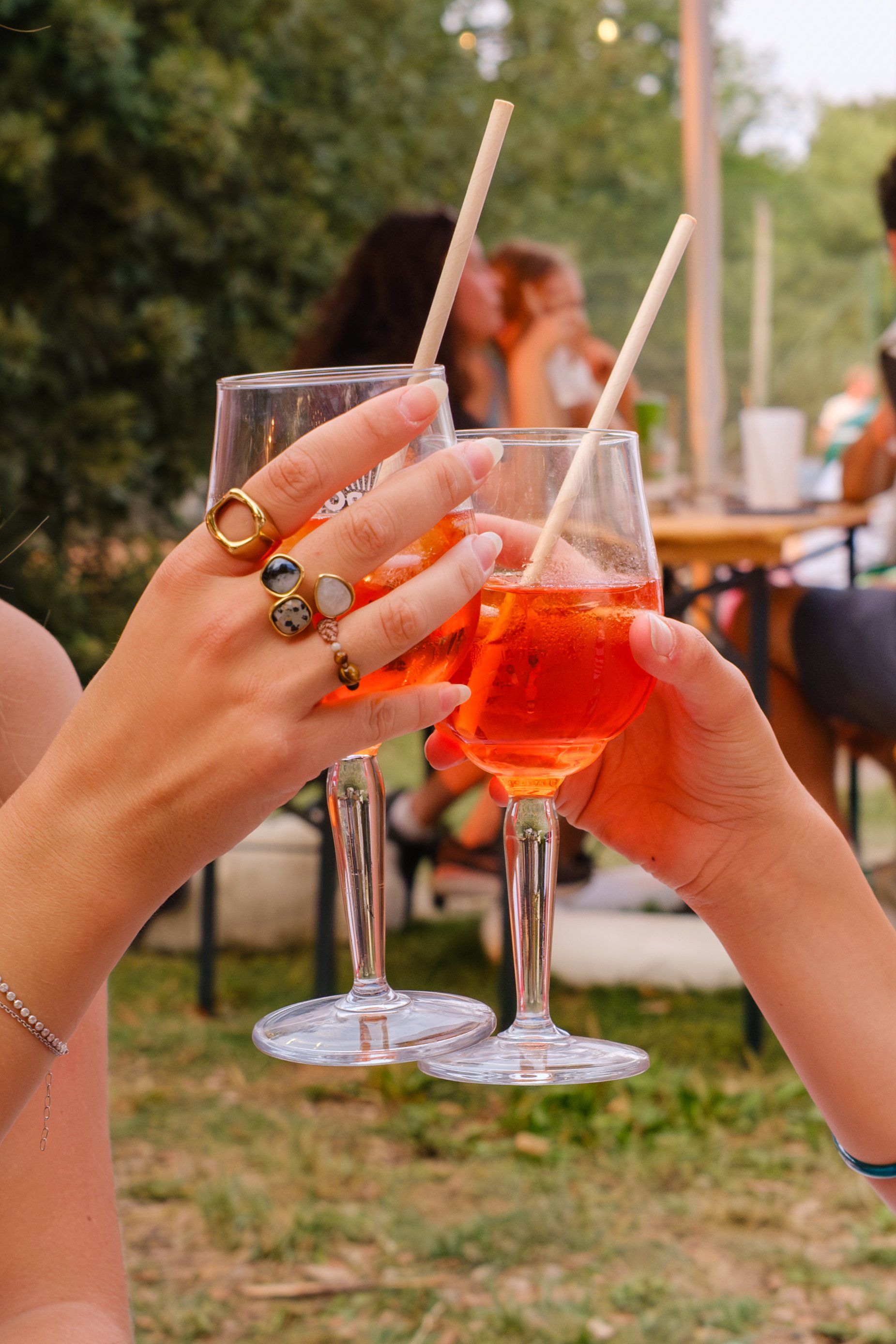 Deux femmes trinquant avec des verres de boisson rose, avec une poignée décorée de plusieurs bagues. En arrière-plan, un groupe de personnes assises dans un espace extérieur.