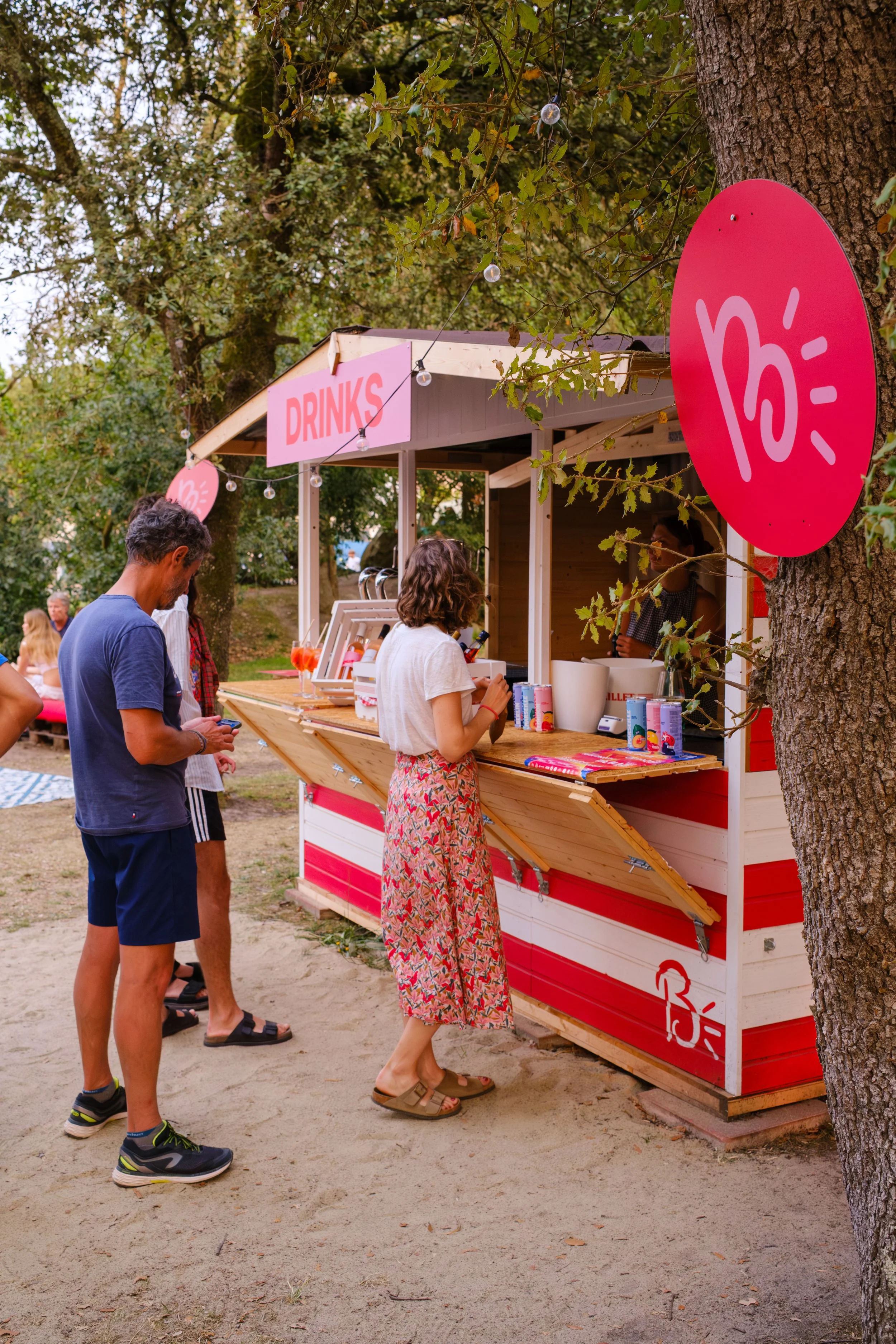 Stand de boissons avec personnes faisant la queue, entouré d'arbres, avec un panneau rose indiquant «DRINKS» et des décorations lumineuses.
