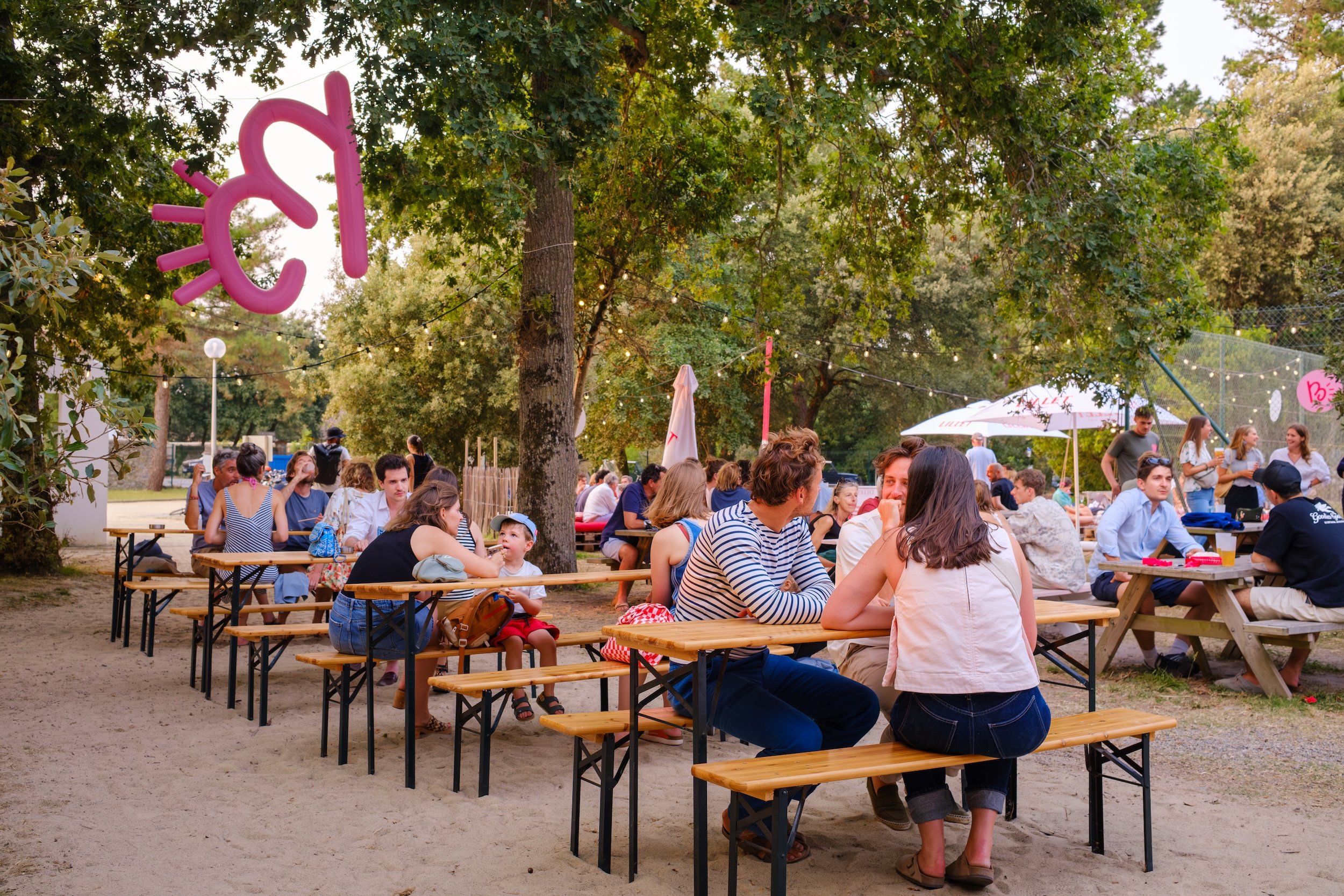 Une scène de fête en plein air avec des personnes assises à des tables en bois sous des arbres, discutant et profitant d'une ambiance conviviale. Des décorations en ampoules et une figure de soleil en ballon rose sont visibles.