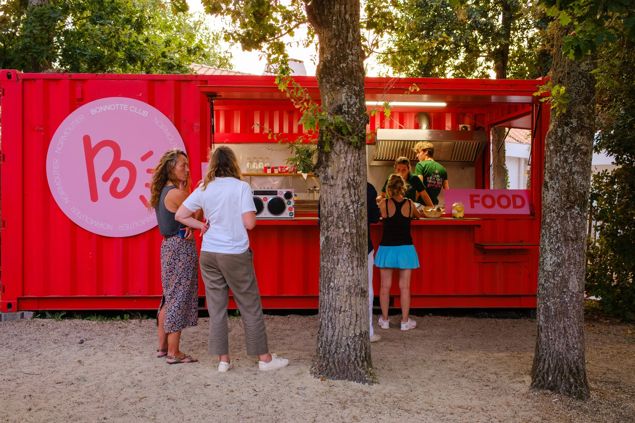 Une file de personnes devant un stand de nourriture rouge en plein air, entouré d'arbres, avec une équipe de cuisine à l'intérieur.
