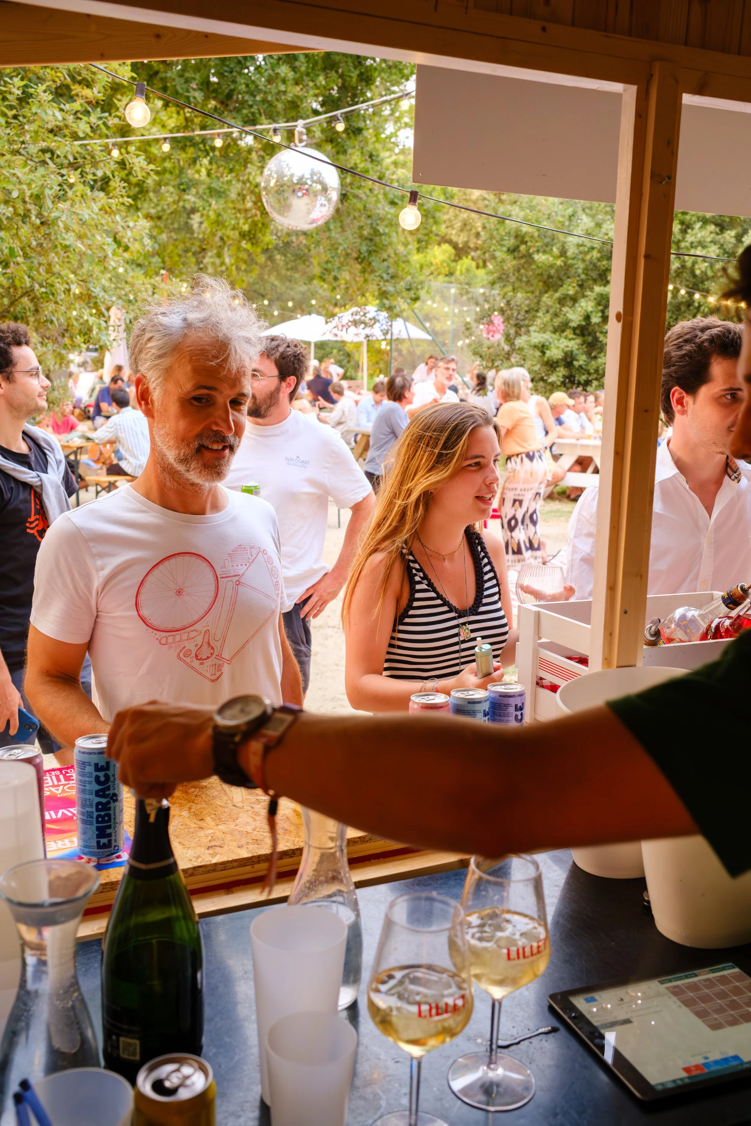 Groupe de personnes à une fête en extérieur, vues à travers une fenêtre de bar ou stand, avec des boissons et décorations lumineuses en arrière-plan.