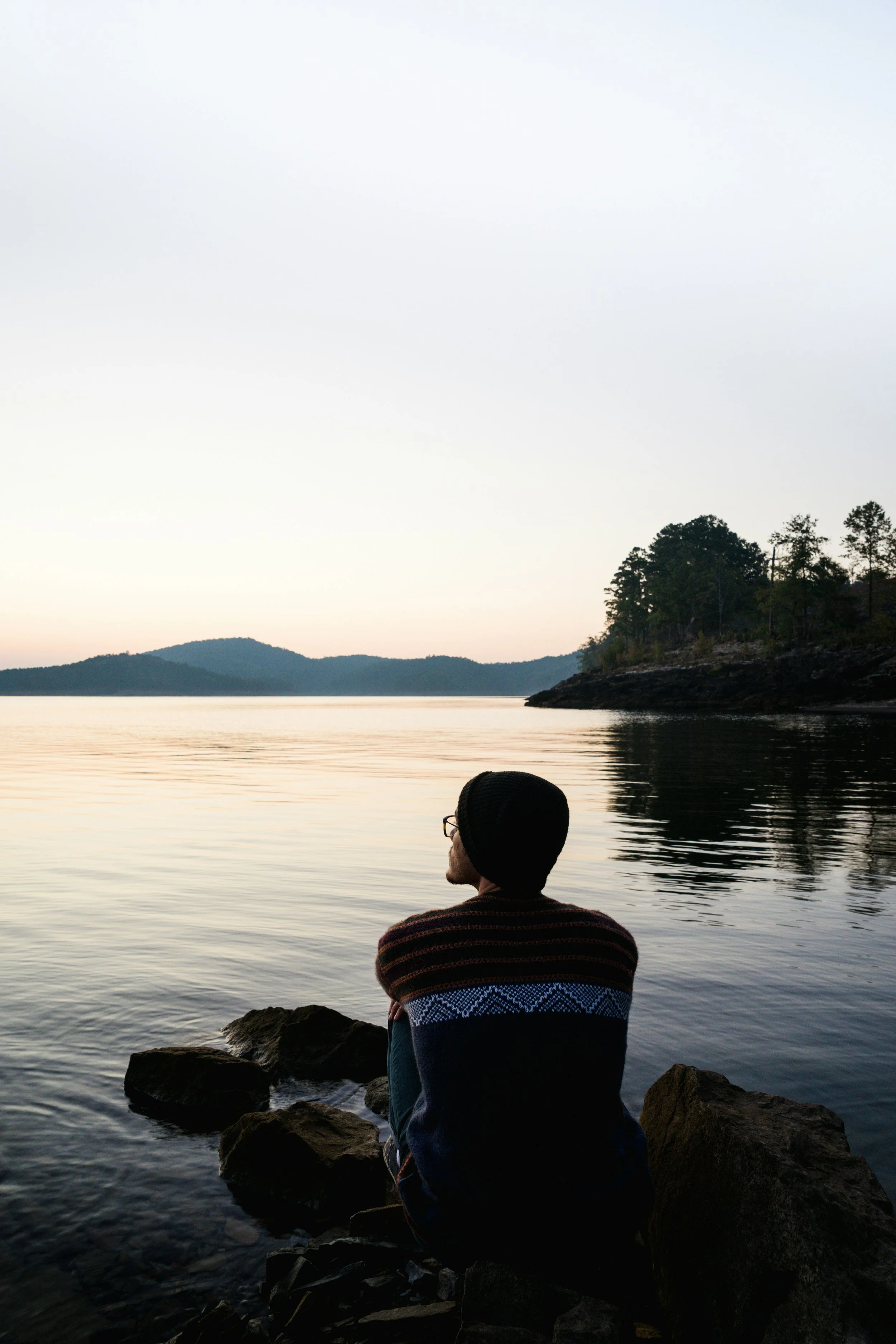 Back view of a person wearing a beanie and warm patterned sweater, sitting peacefully on lakeside rocks at dawn and looking out over still, calm water toward distant misty hills.