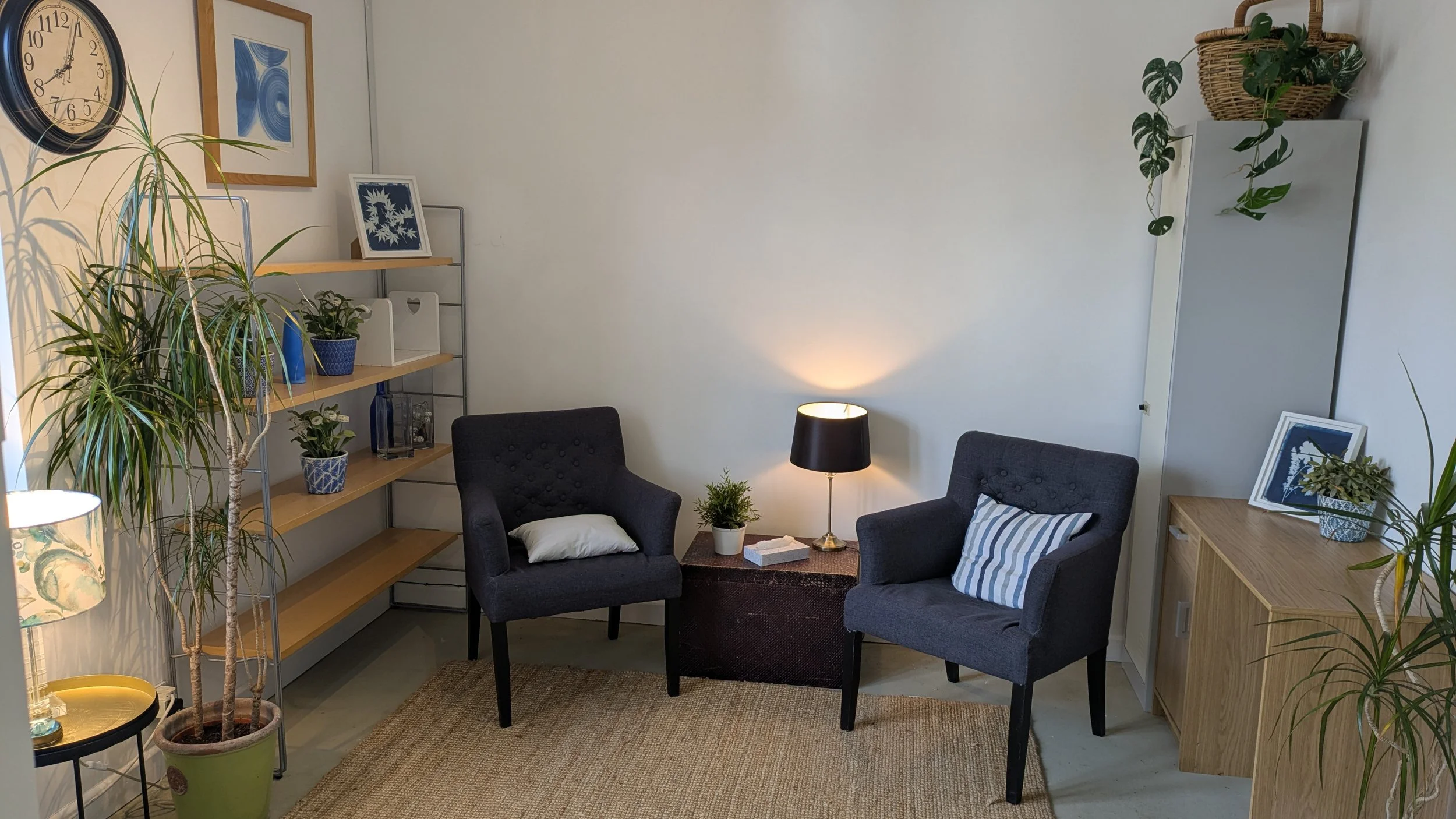 A calm and inviting therapy room at Beanbag Natural Health in Witney, featuring two comfortable blue armchairs, natural wood shelving with plants, and warm lamp lighting.