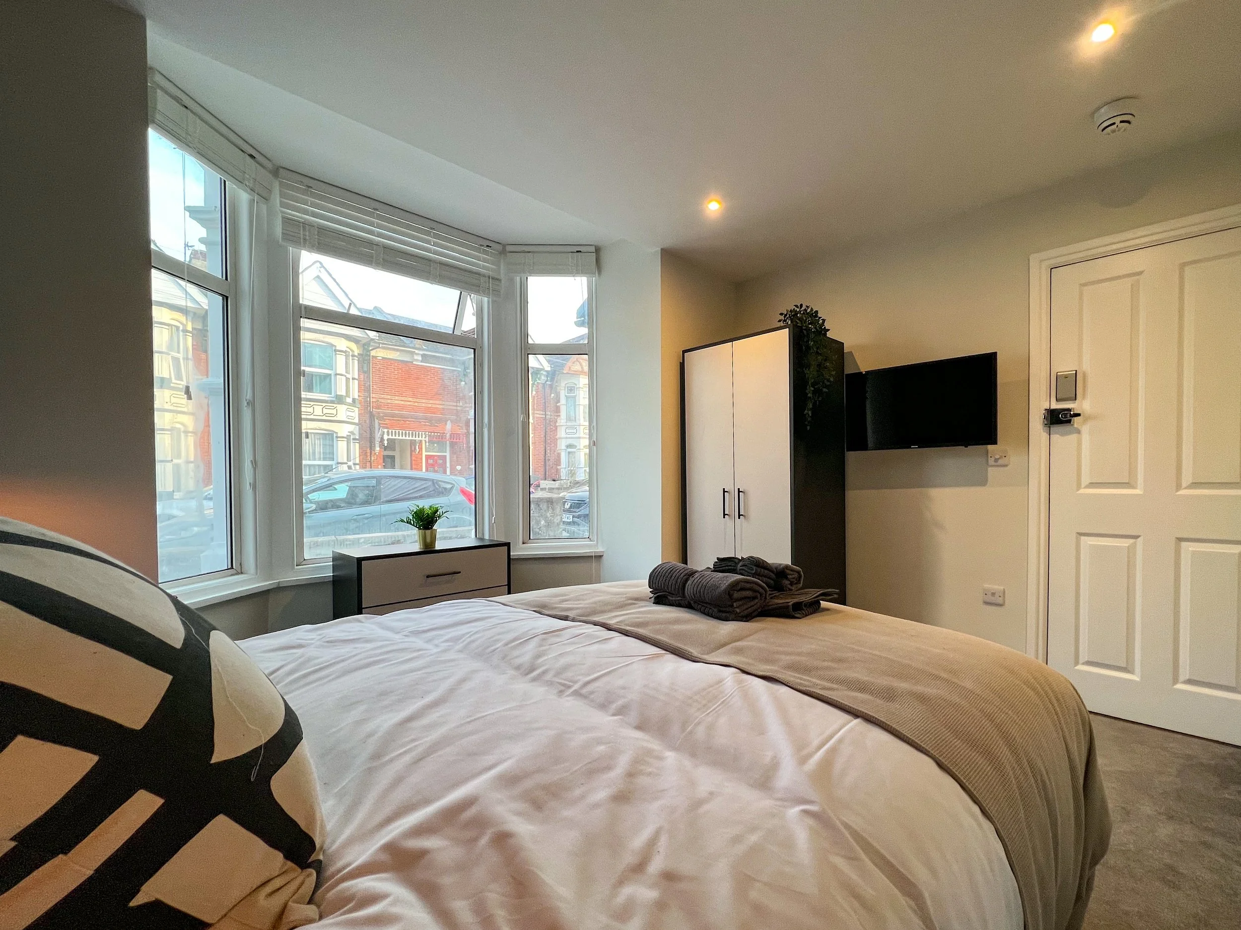 A cozy bedroom with large bay windows, a bed with beige and white bedding, stacked towels on the bed, a black wardrobe, a mounted flat-screen TV, a small black and white dresser with a potted plant, and a white door. There are blinds on the windows a