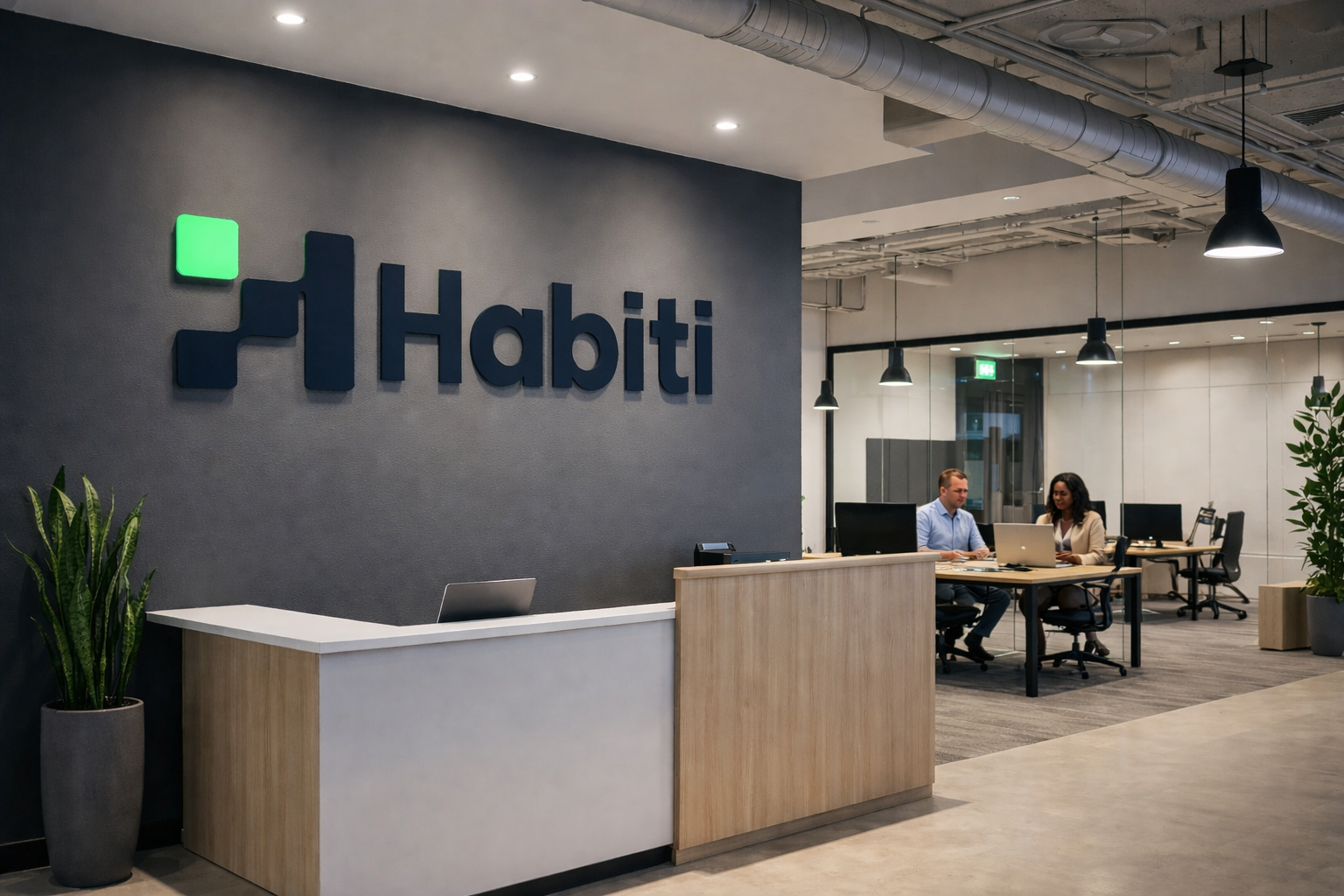 Modern office reception area with a dark grey wall featuring the Habiti logo in blue and a small green square. There is a white and wooden reception desk with a laptop and a plant nearby. In the background, two people are seated at a table working on laptops, with black pendant lights hanging above and glass walls separating the office space.
