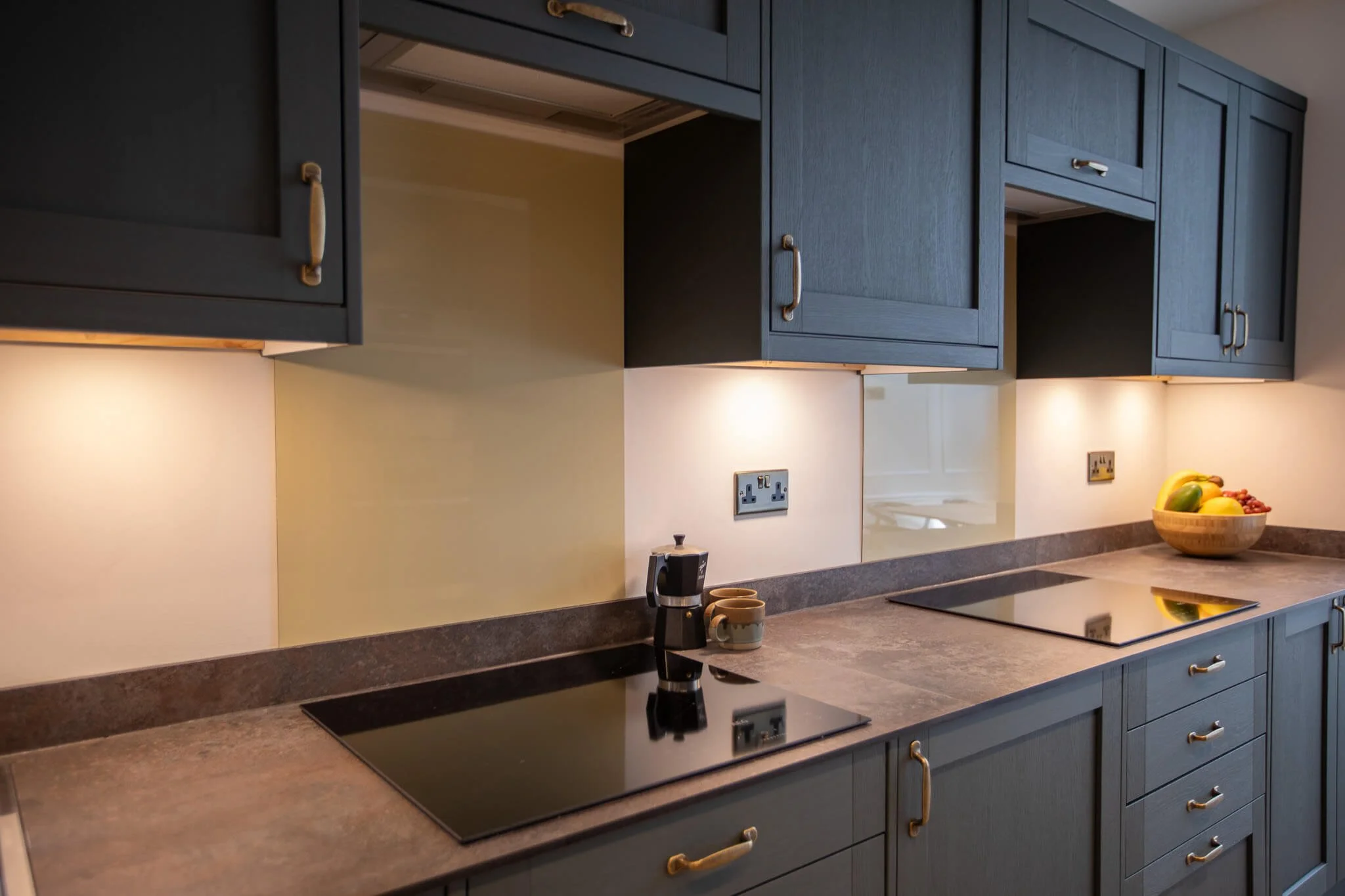 Kitchen with dark blue cabinets, beige walls, gray countertop, and built-in electric stovetops. There is a bowl of fruit on the right, a coffee machine and two mugs near the center, and electrical outlets on the wall.
