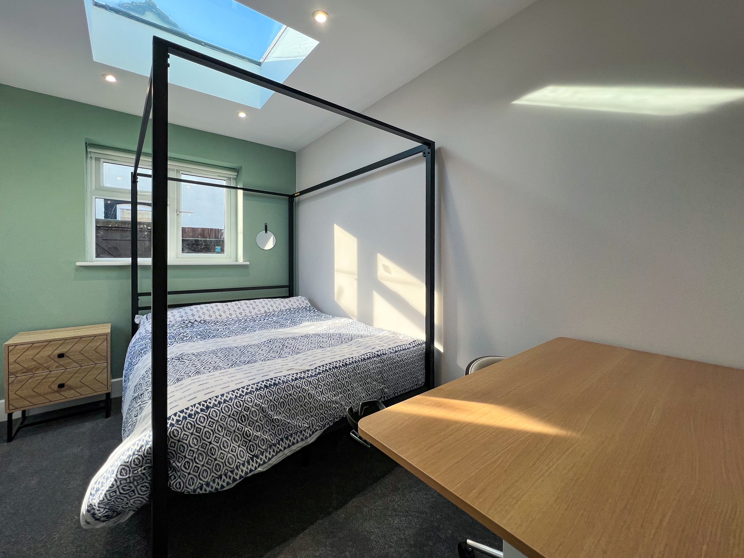 A bedroom with a bed under a black metal frame, a wooden nightstand, a window, and a ceiling skylight with some sunlight coming in.