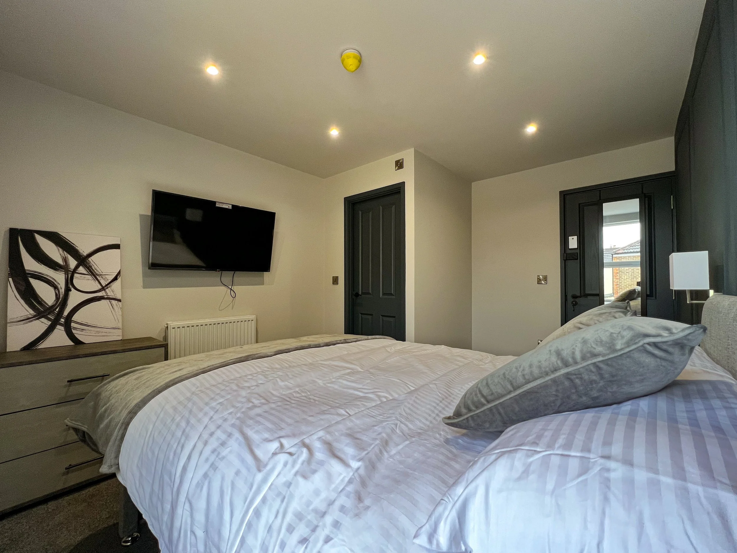 A bedroom with a bed, pillows, dresser, wall-mounted TV, and a door leading outside. The room has white walls, a ceiling with multiple recessed lights, and black door frames.