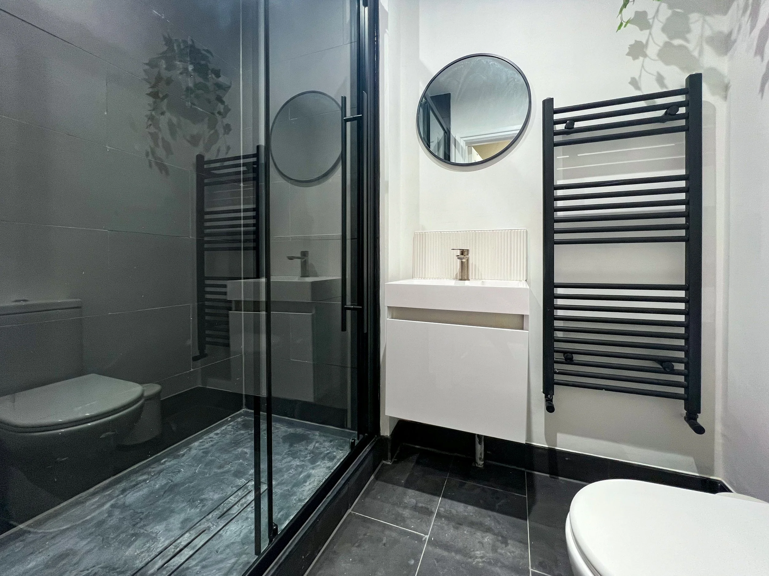 Modern bathroom with gray tiled shower, black ladder towel rack, round mirror, white vanity with sink, and black floor tiles.