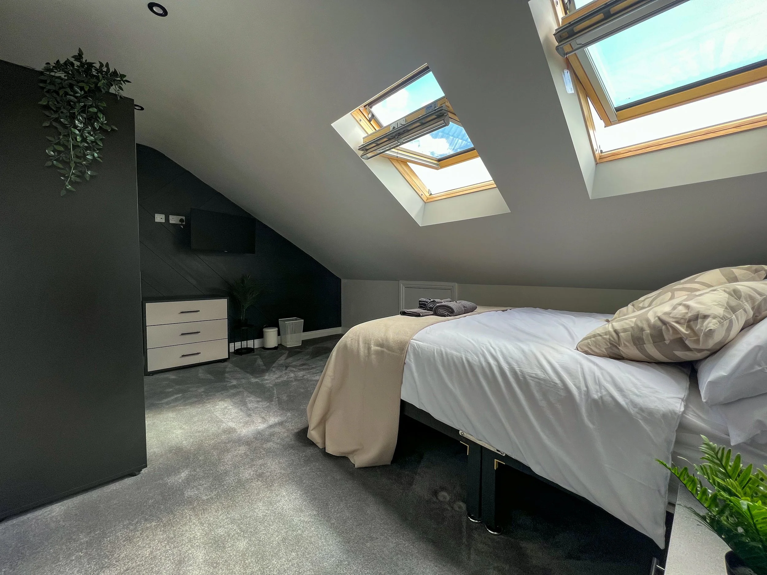A modern, minimalist bedroom with a sloped ceiling and two skylight windows letting in natural light. The bed has white sheets and beige pillows, with a beige blanket folded at the foot. There is a black and white dresser, plants, and a small televis