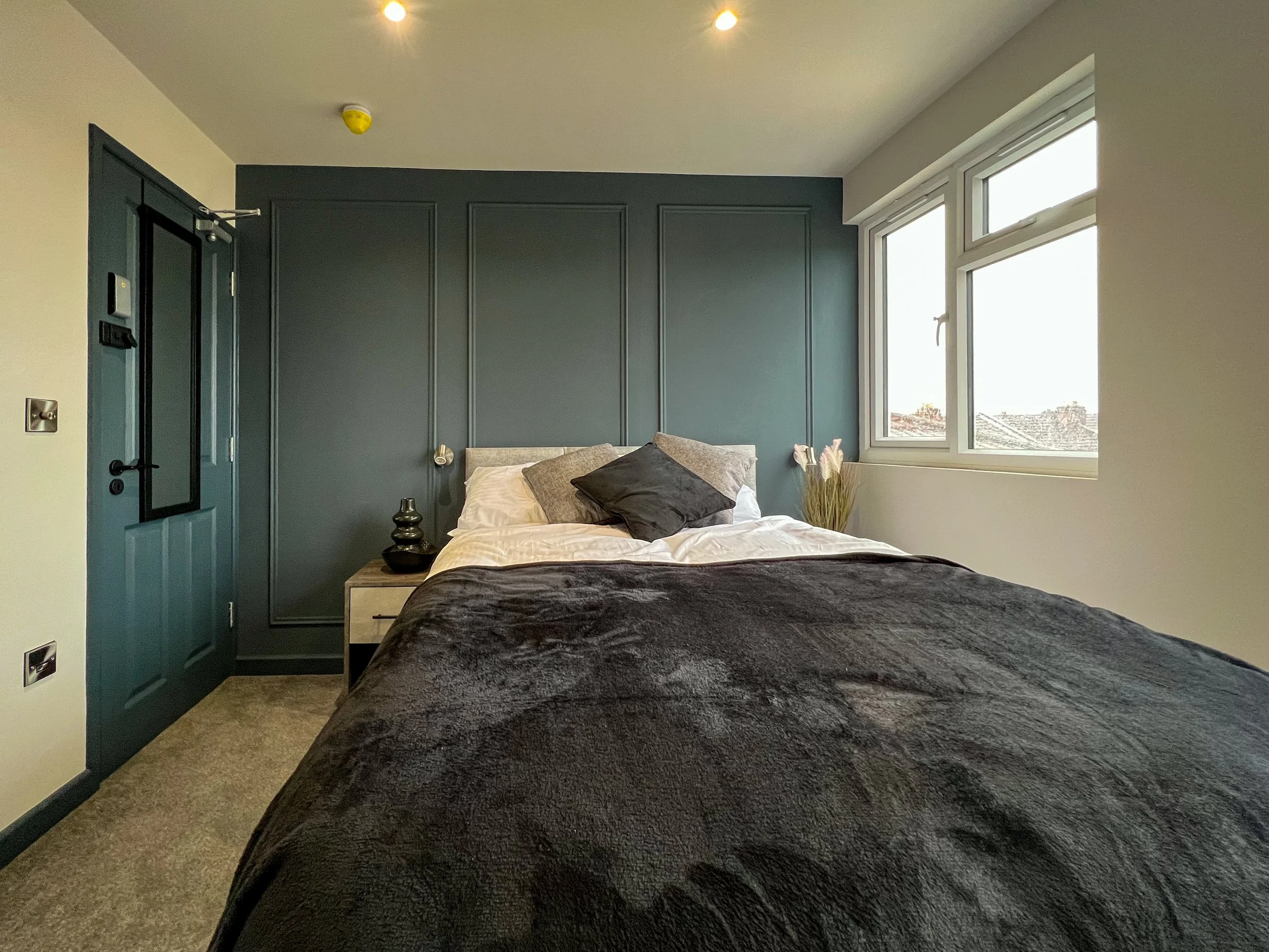 A bedroom with dark teal accented wall and door, light-colored bed with grey, black, and beige pillows, small bedside table with decorative objects, and a window showing rooftops outside.