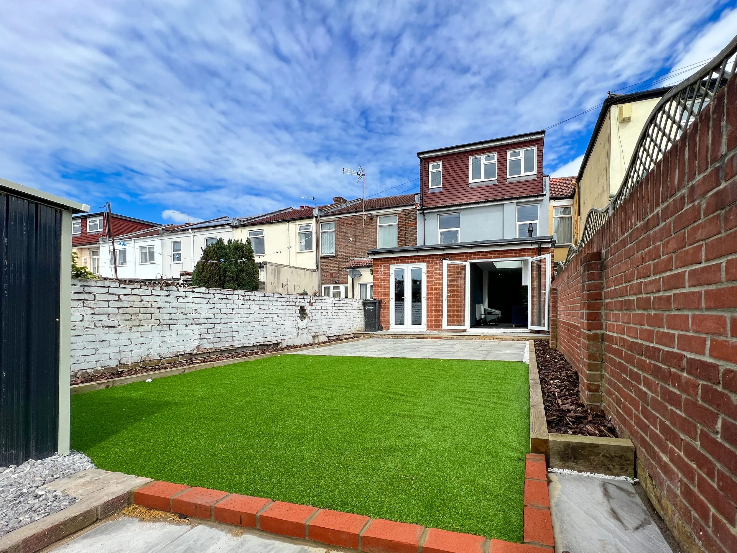 View of a backyard with a lawn and a patio, enclosed by brick and stone walls, with a residential house and neighboring houses in the background under a partly cloudy sky.
