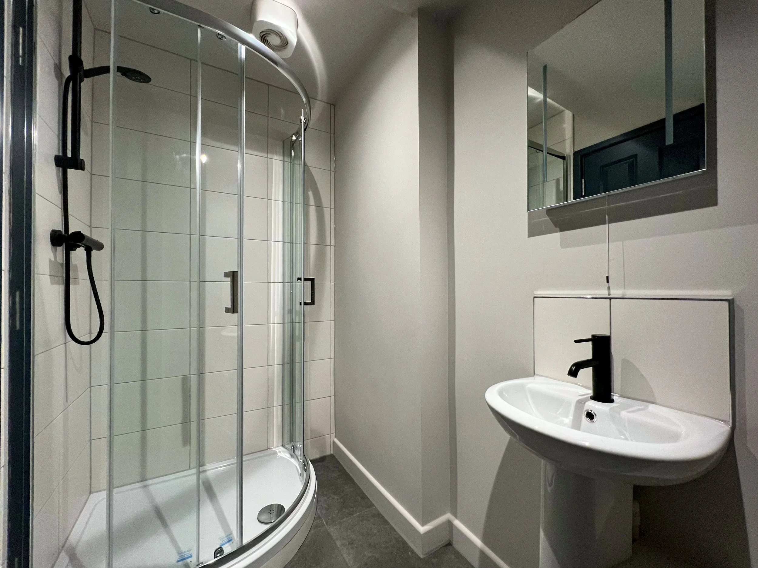 A modern bathroom featuring a glass-enclosed curved shower with a black showerhead and handle, a small white sink with a black faucet, a rectangular mirror above the sink, and a window with a pull-down shade.