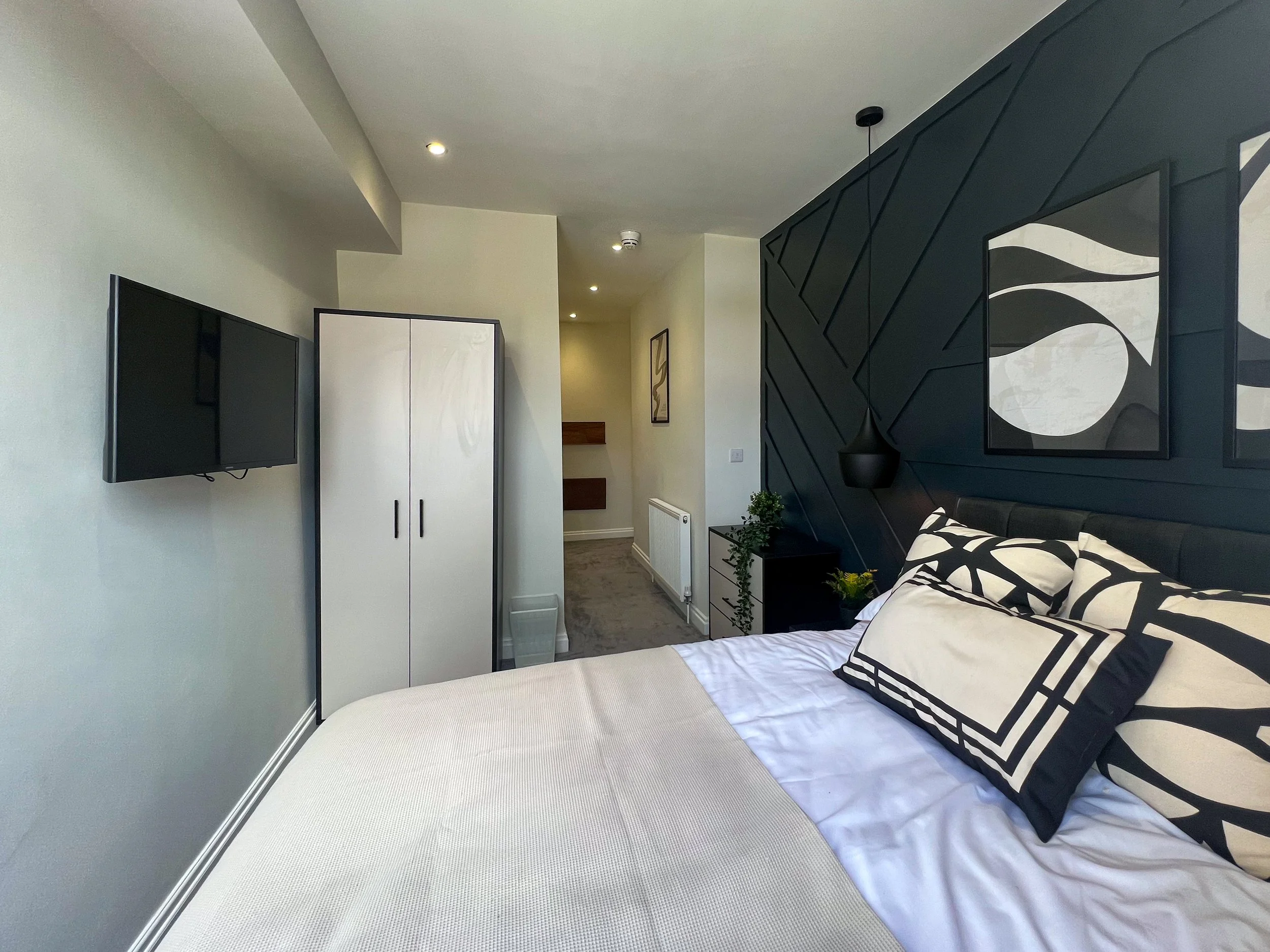 A modern bedroom with a large bed, black and white decorative pillows, a black accent wall with artwork, a wall-mounted television, a white wardrobe, a small black nightstand with plants, and a hallway in the background.