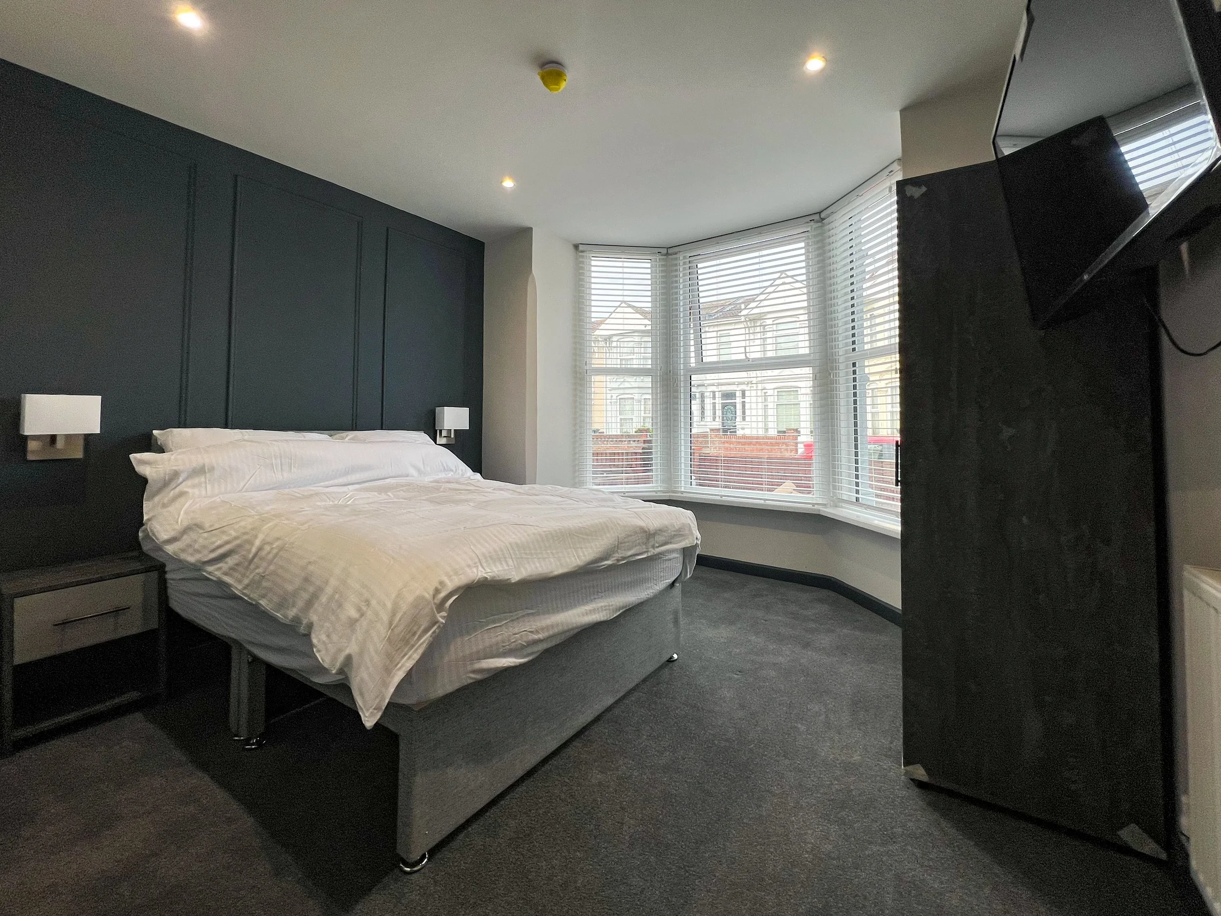 A bedroom with a bed, nightstand, dark-colored headboard wall, and large bay window with white blinds.