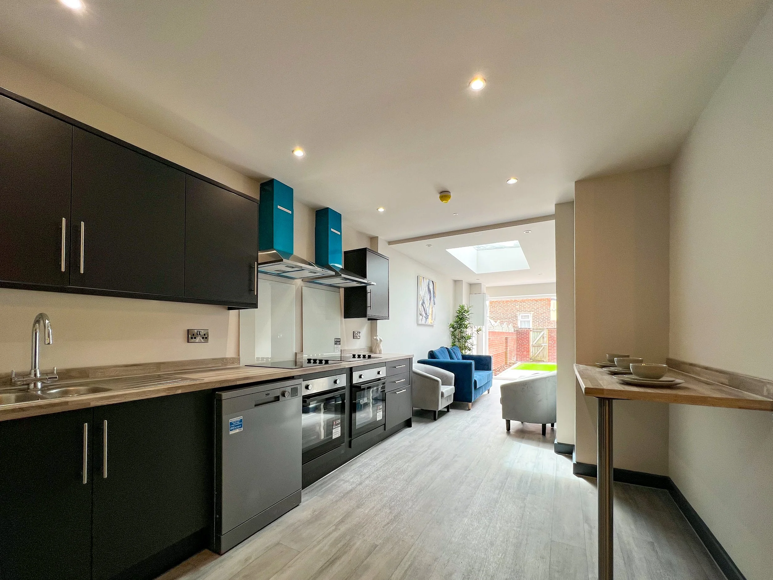 Modern open-plan kitchen and living space with black cabinets, stainless steel appliances, seating area with chairs and sofa, and a view of a backyard through sliding glass doors.