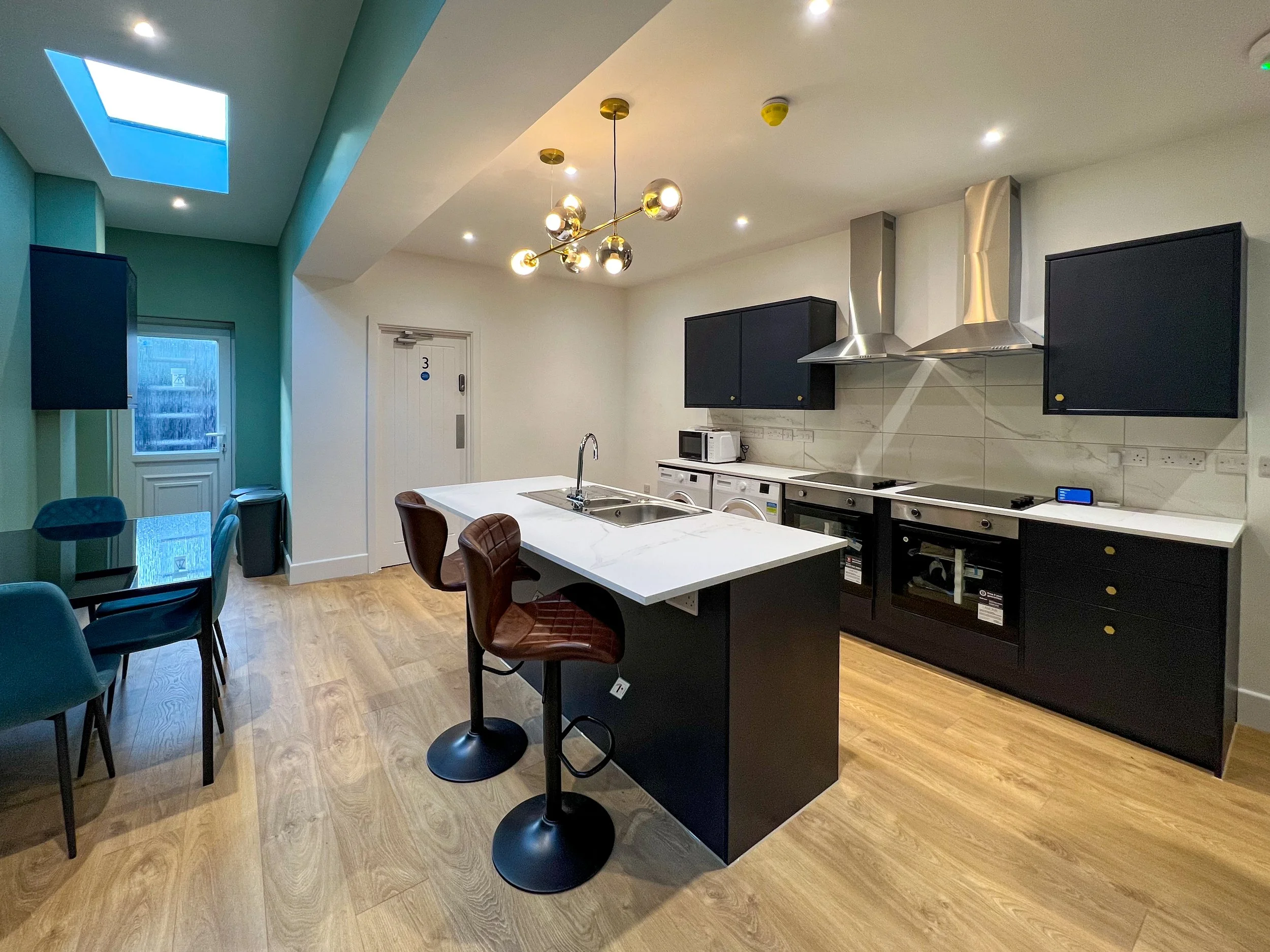 Modern kitchen with black cabinetry, marble countertop island, stainless steel range and range hoods, and a dining area with teal and blue chairs, hardwood flooring, and a skylight.