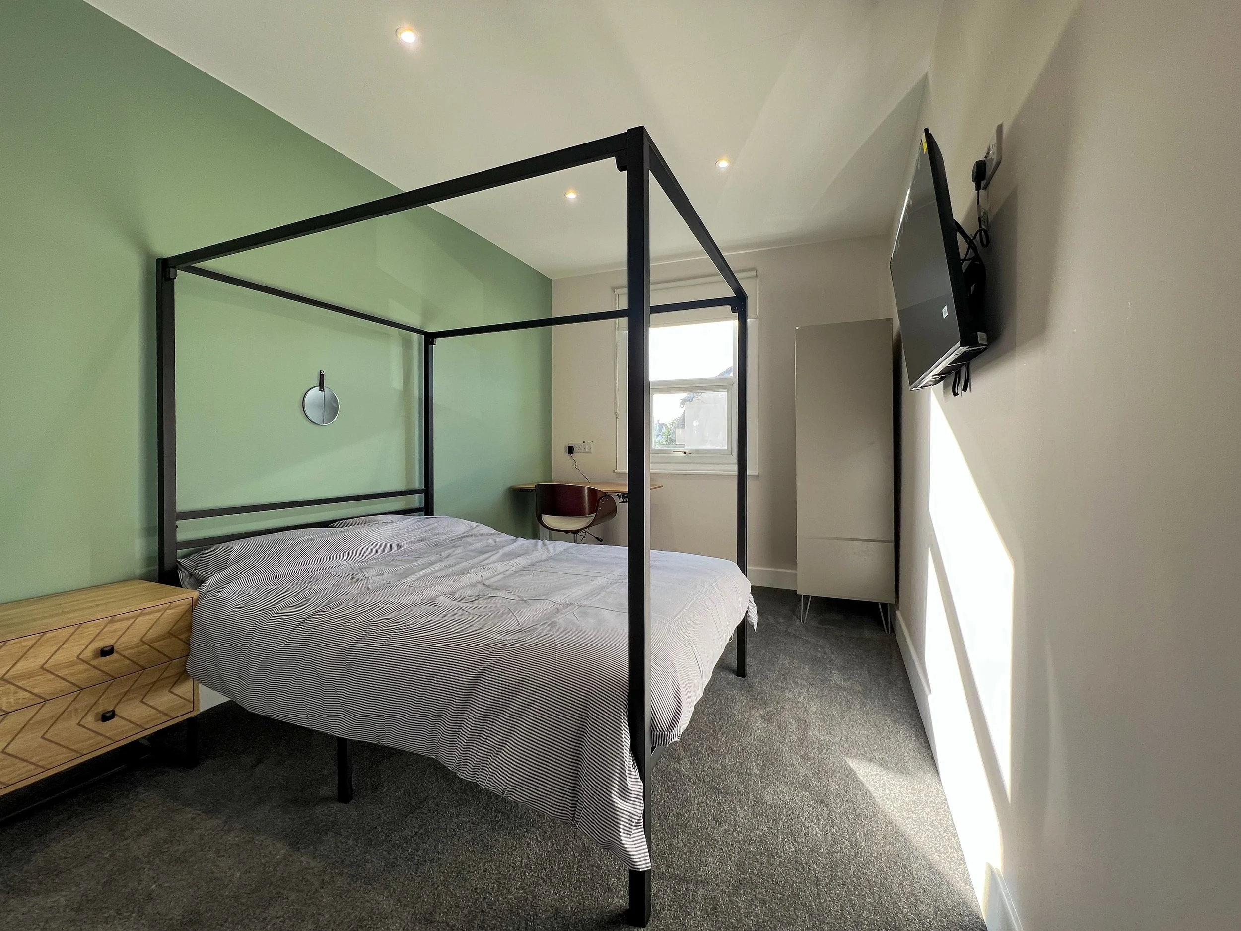 Modern bedroom with a black metal canopy bed, grey striped bedding, a wooden nightstand, a small desk with a chair, a wall-mounted TV, and a window letting in natural light, with a green accent wall and a neutral-colored wall.