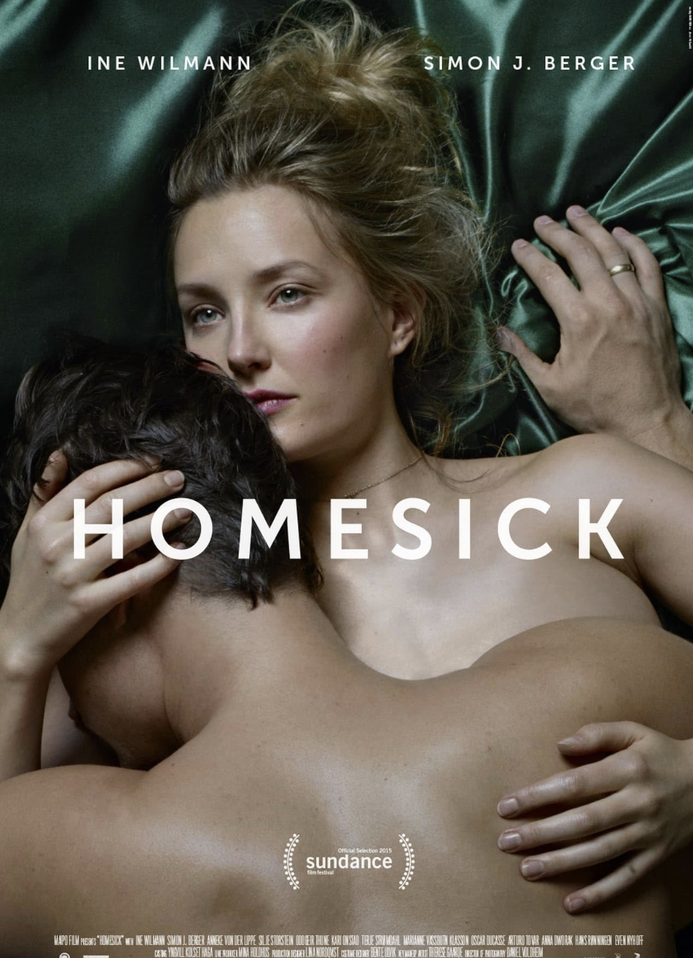 A woman with blond hair lying on a green satin sheet, embracing a shirtless man with dark hair. The woman's hand is resting on the man's head, and her face is looking intently at something. The text on the image is the title "HOMESICK" and the names "INE WILMANN" and "SIMON J. BERGER."