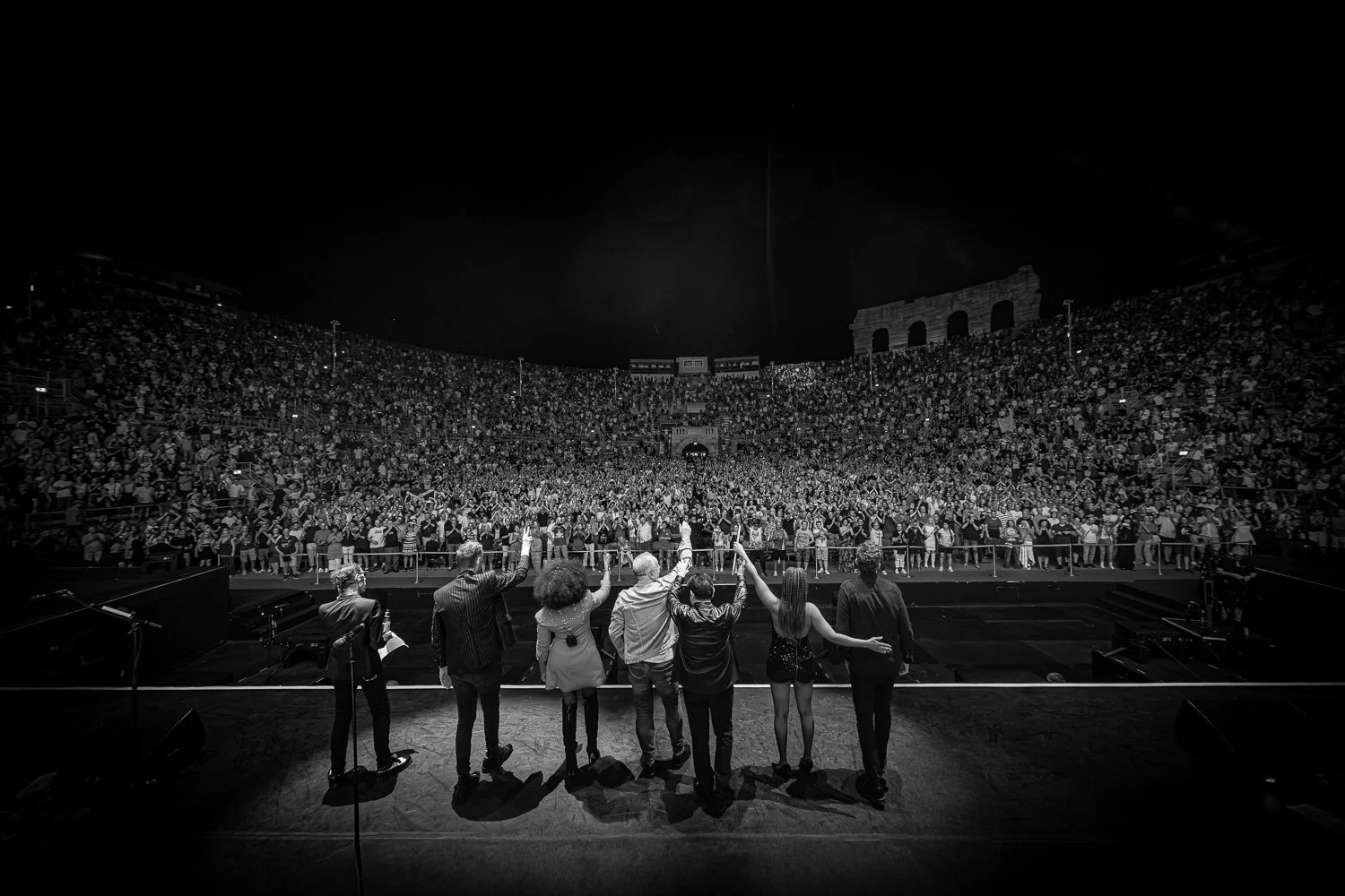 A black and white photo of an audience at a large indoor concert, with performers on stage waving to the crowd.