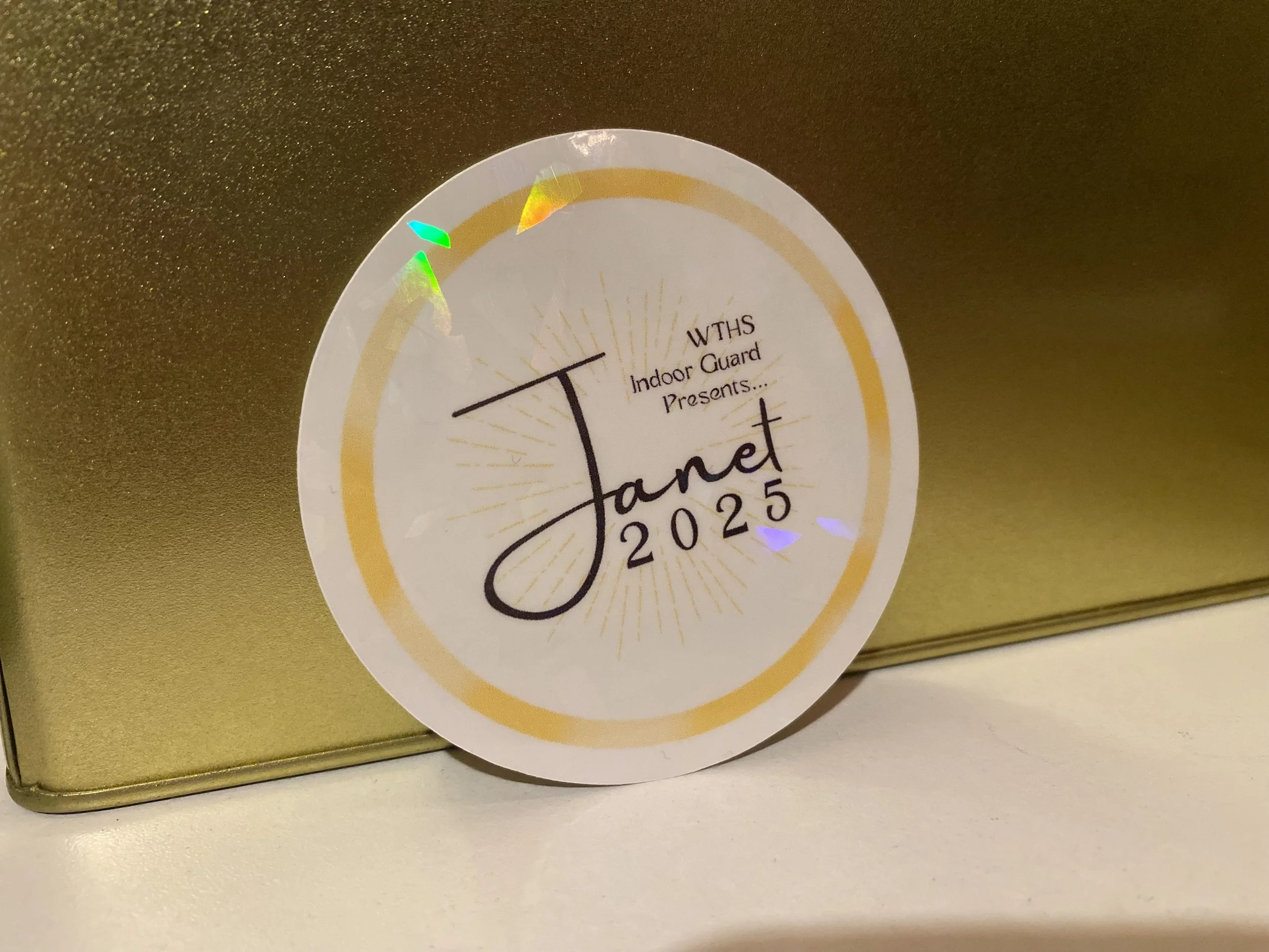 A circular sticker with the text "WTHS Indoor Guard Presents... Janet 2025" on a reflective gold surface.
