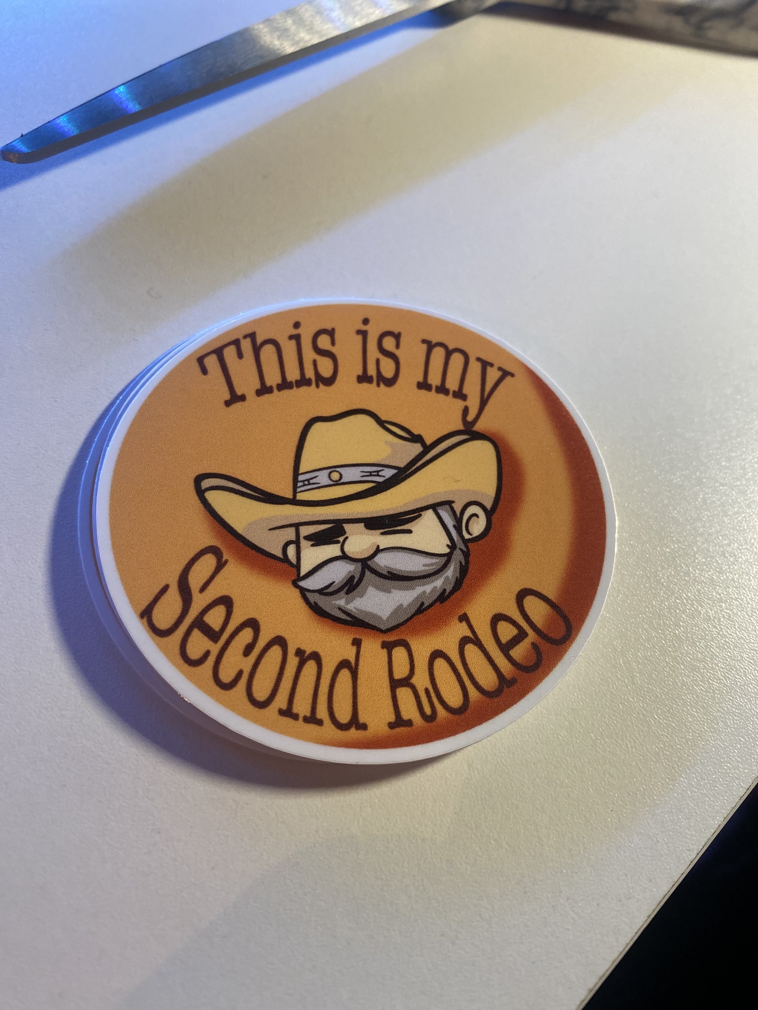 A sticker with a cartoon man wearing a cowboy hat and a beard, with the text 'This is my Second Rodeo' on an orange background.