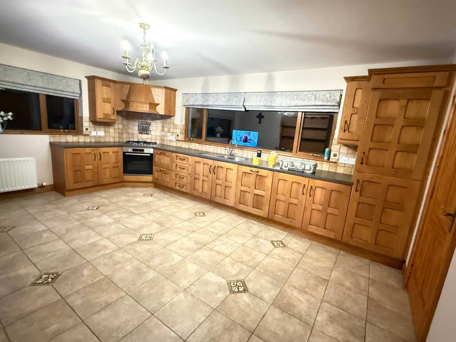 A spacious kitchen with wooden cabinets, tiled floor, large windows, and a chandelier. The countertop has some items including cleaning supplies, and the kitchen features a built-in oven and stove with a vent hood.