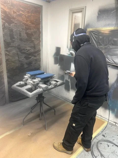 A man wearing a mask, headphones, and black clothing is spray painting a wall in a room. The room contains a small round table with some objects on it, including a few closed blue books or binders. The wall and nearby surface are covered with black s