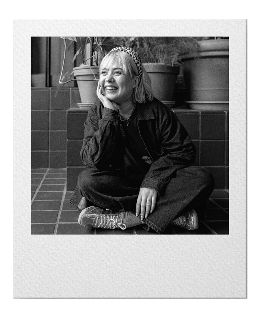 Black and white photo of a woman sitting on a tiled floor, smiling and laughing, resting her chin on her hand, with potted plants in the background.