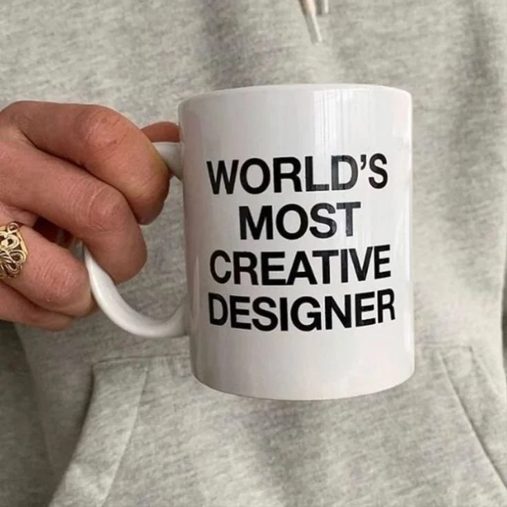 White coffee mug with black text that reads 'World's Most Creative Designer', being held by a hand with a gold ring.