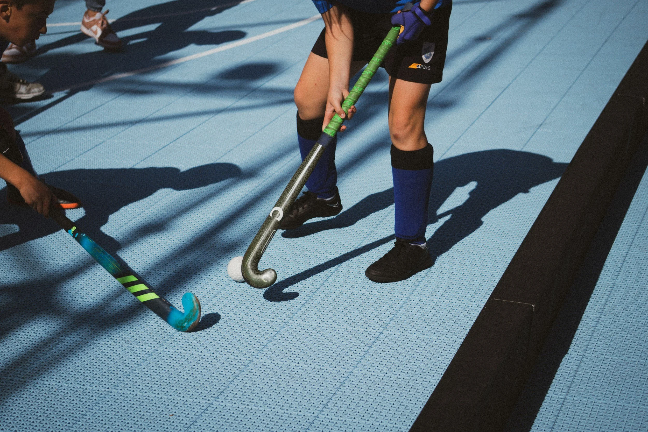 A person dressed in black sports attire and blue knee-high socks is playing indoor hockey on a blue textured surface, using a hockey stick to hit a white ball. They are accompanied by another person, partly visible, also with a hockey stick, and other players are in the background.