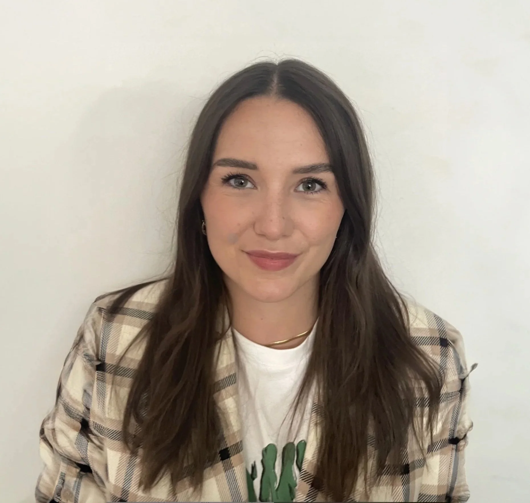 Young woman with long brown hair, light makeup, wearing a plaid blazer over a white T-shirt with green graphic, standing against a white wall.