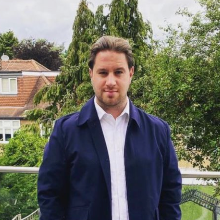 A young man with light skin and brown hair standing outdoors on a balcony, wearing a white shirt and a navy blue jacket, with trees and houses in the background.