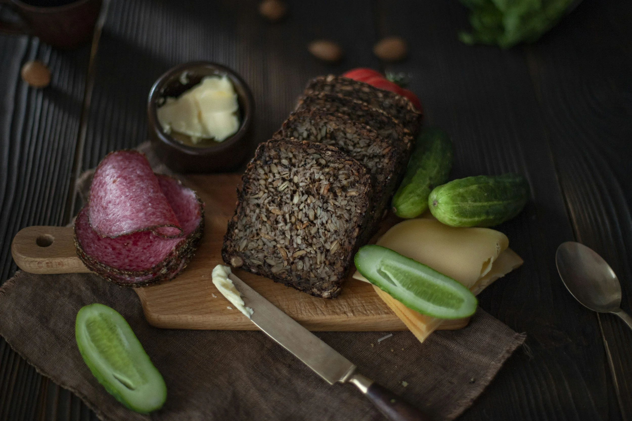 Seed bread with butter, salami and pickles on a wooden serving board, against a dark linen covered, wooden table