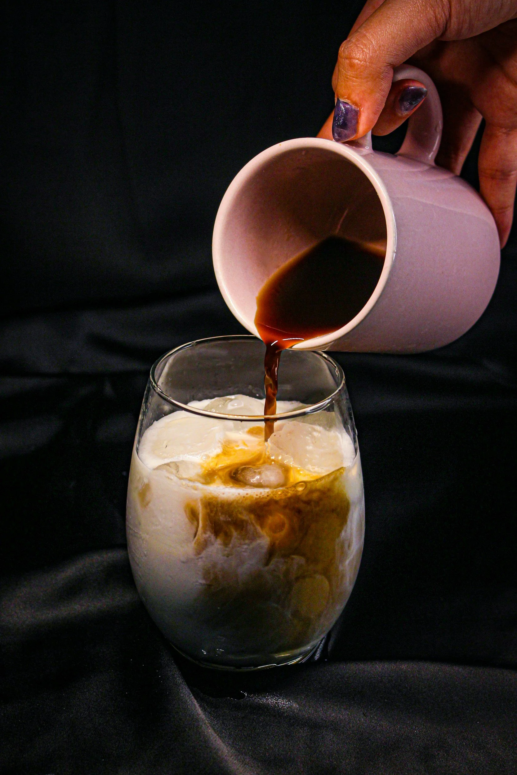 A hand pouring coffee into a glass holding a milk and ice mixture