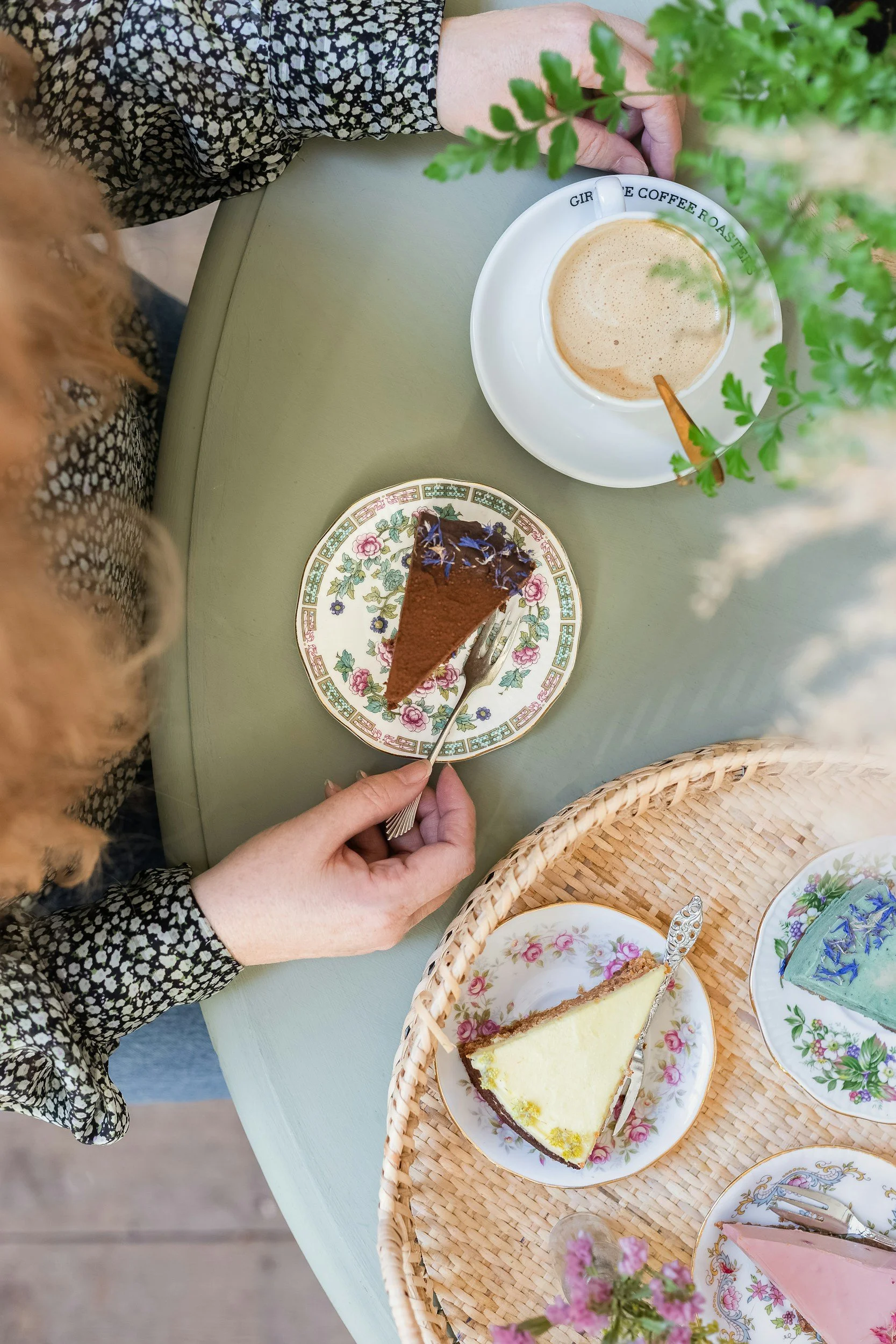 A woman with red hair eating chocolate cake, with a coffee beside here, and an addition tray of cakes on a green table