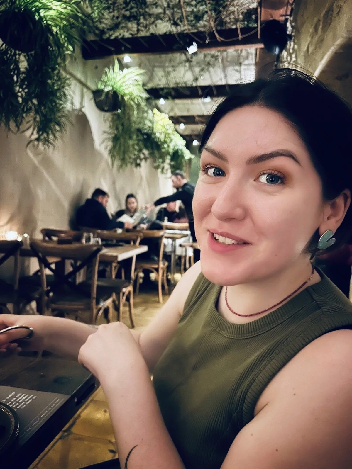 A woman with dark hair, wearing a green sleeveless top and earrings, sitting at a restaurant table with other diners and green plants in the background.