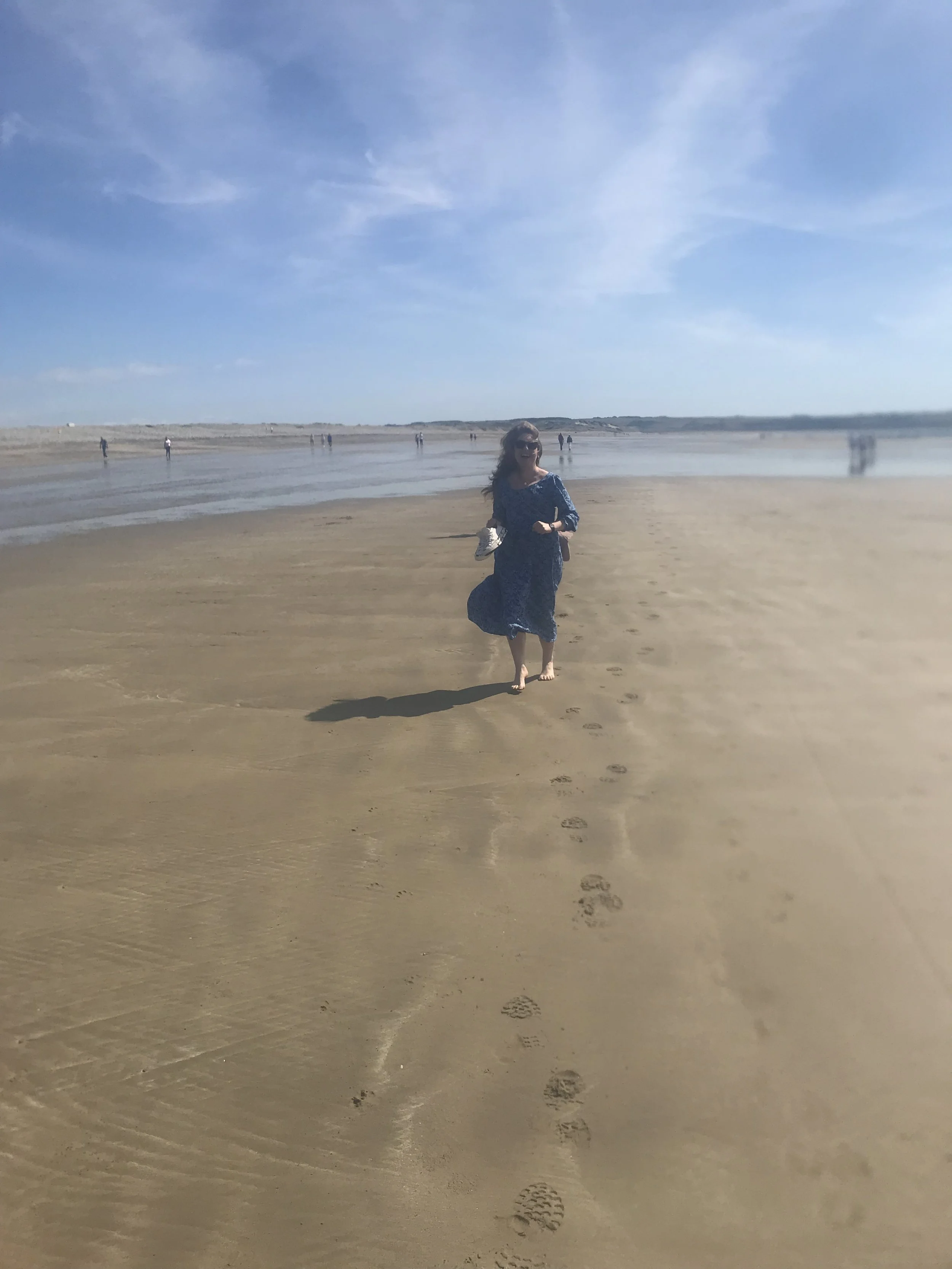 A woman in a blue dress holding white shoes running backwards on a beach