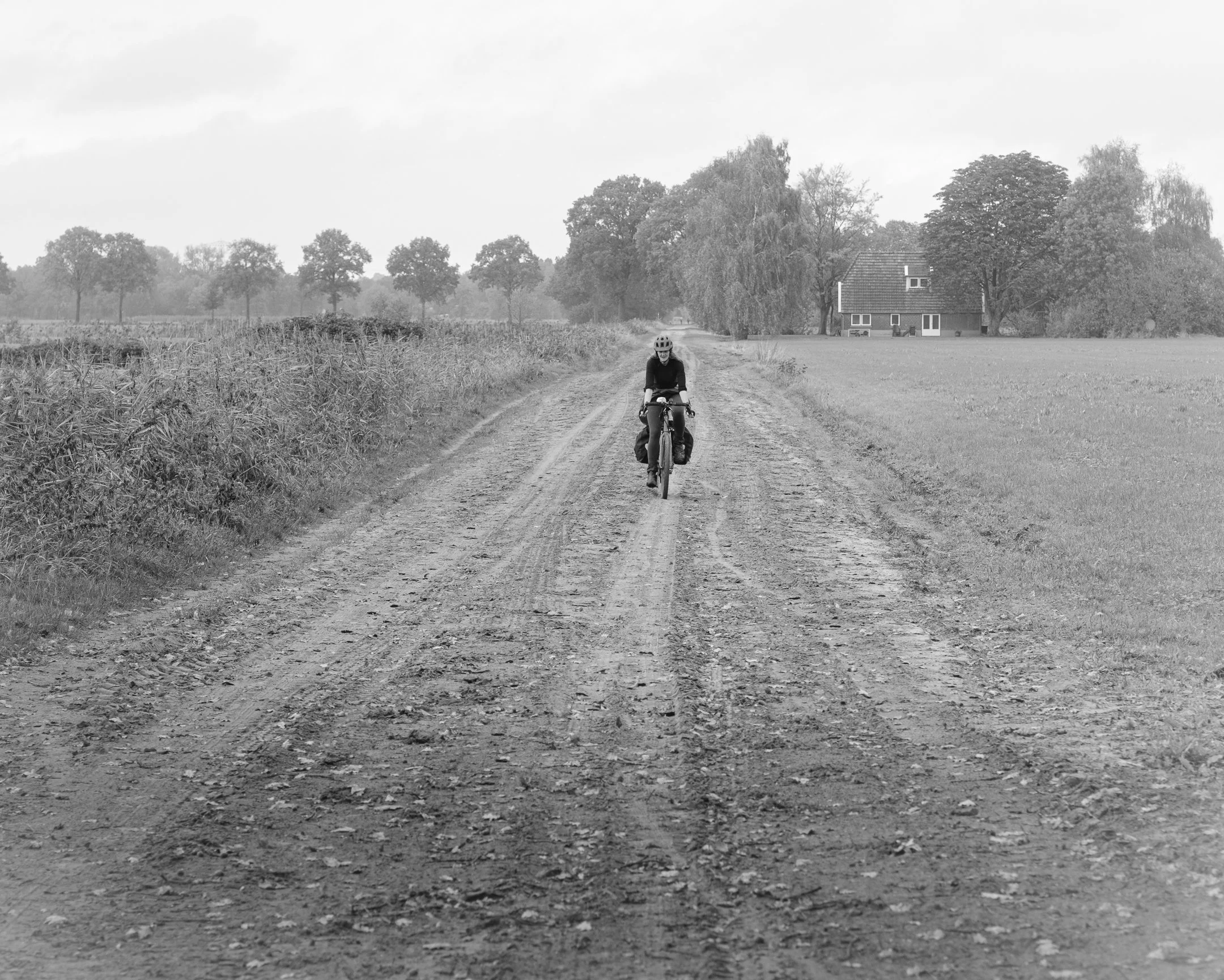 Een persoon fietst op een gravelweg in een landelijke omgeving met grasvelden en bomen, met een huis op de achtergrond.