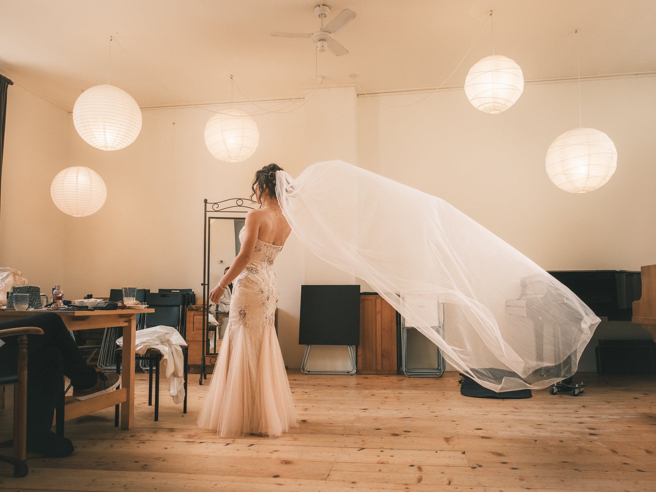 A bride in a strapless gown with lace detailing stands in a room with hanging paper lanterns, her veil flowing behind her.