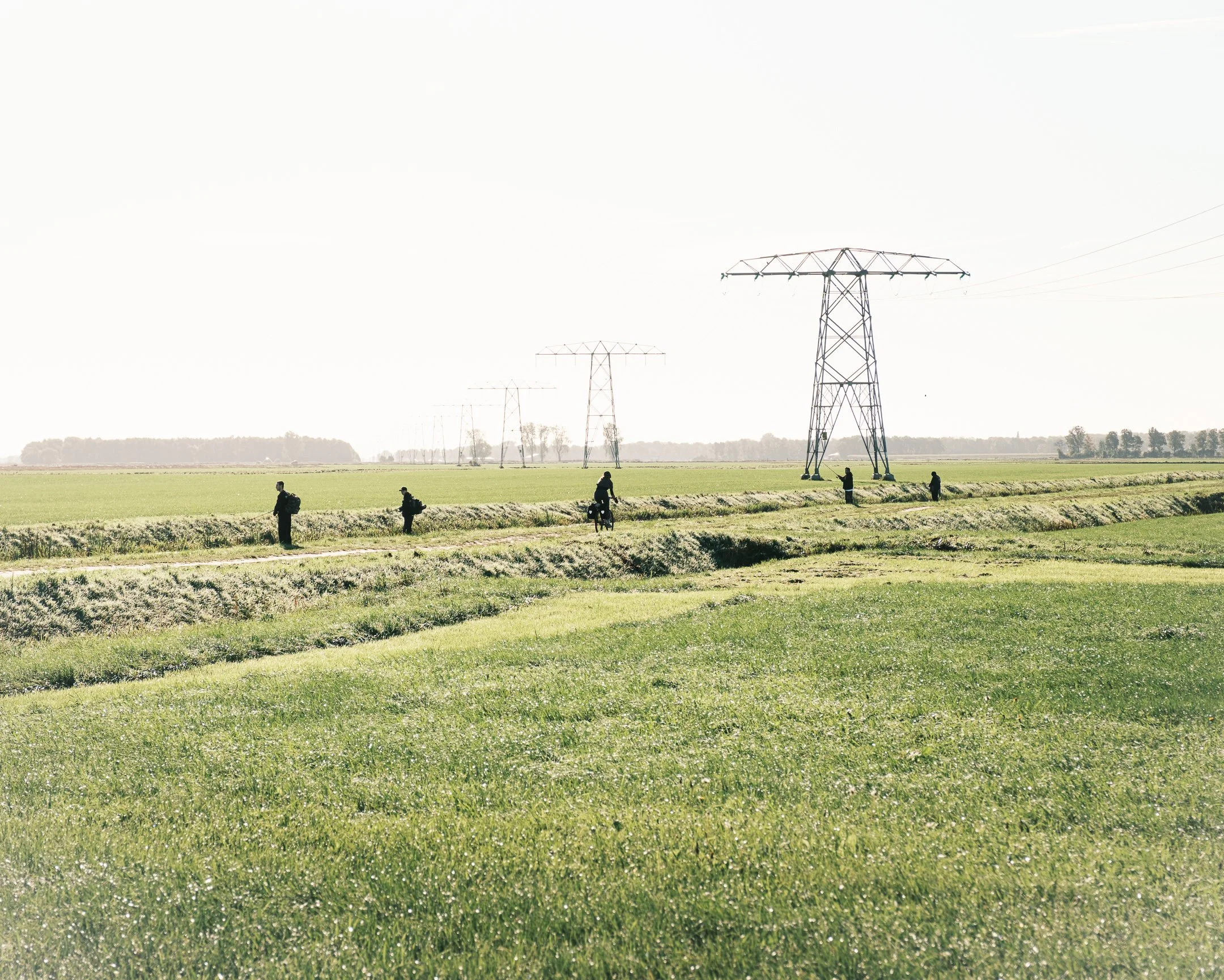 Vijf mensen lopen en fietsen door een uitgestrekt groen landschap met elektriciteitspalen op de achtergrond.