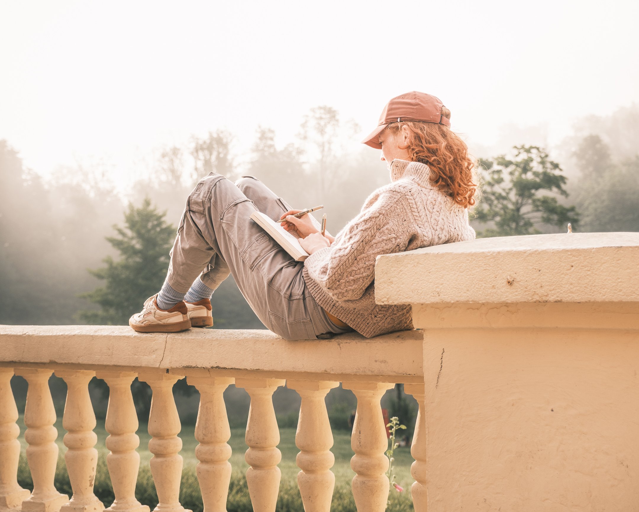 Vrouw met rood haar in een beige trui, grijze broek en een roze pet, zit op een muur en schrijft in een notitieboekje, met een bos in de achtergrond.