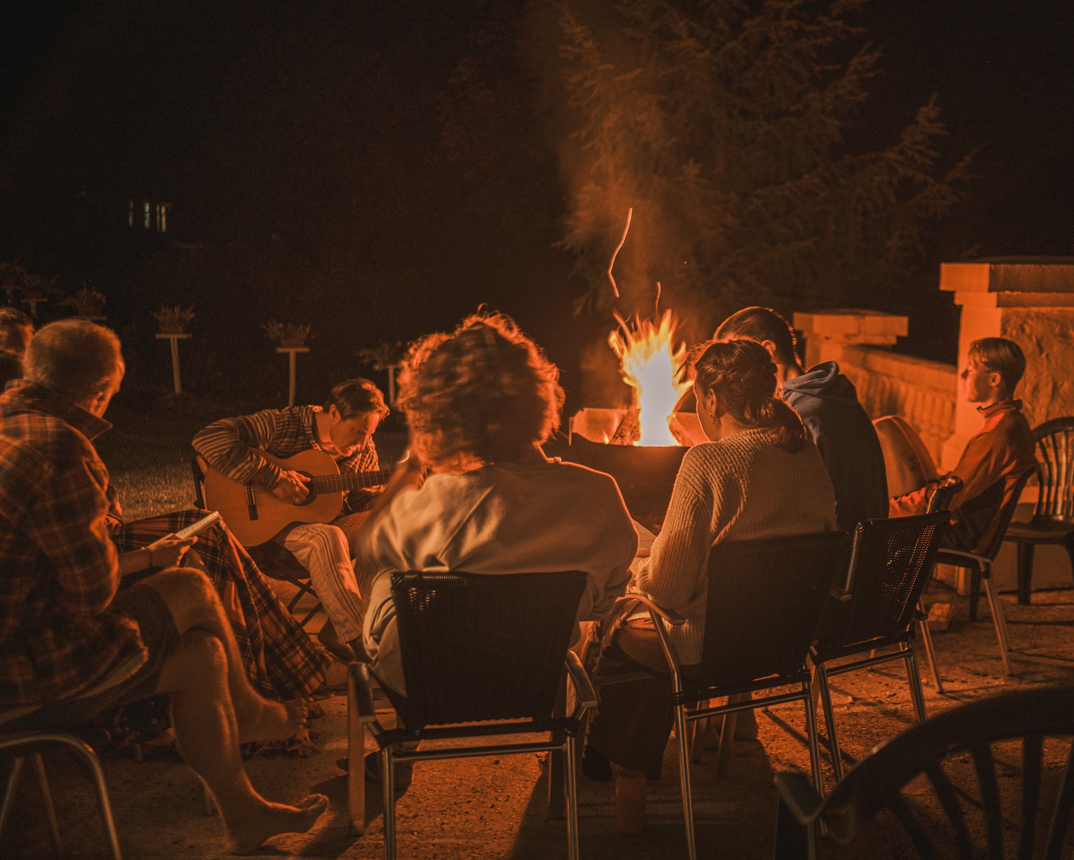 Mensen zitten rondom een kampvuur in de nacht, sommige spelen gitaar, anderen genieten van het samenzijn.
