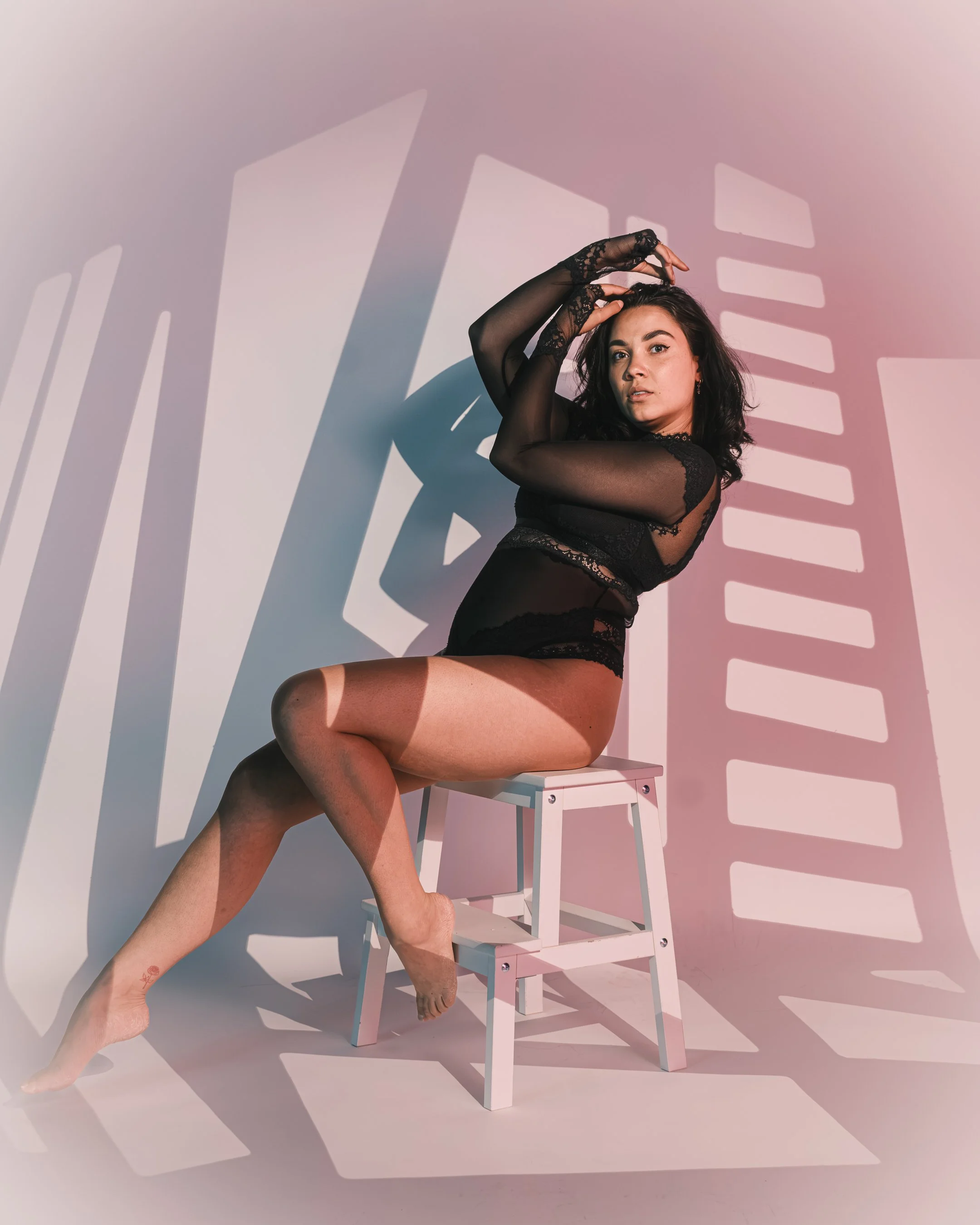 Een vrouw in zwart lace lingerie poseert zittend op een witte stoel, met haar handen boven haar hoofd, terwijl haar schaduw geprojecteerd wordt op de achtergrond.